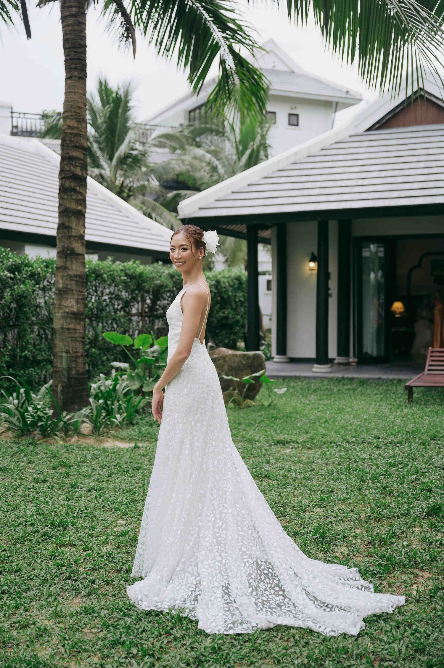 Bride in elegant white lace wedding gown with train poses in lush tropical garden beside palm trees.