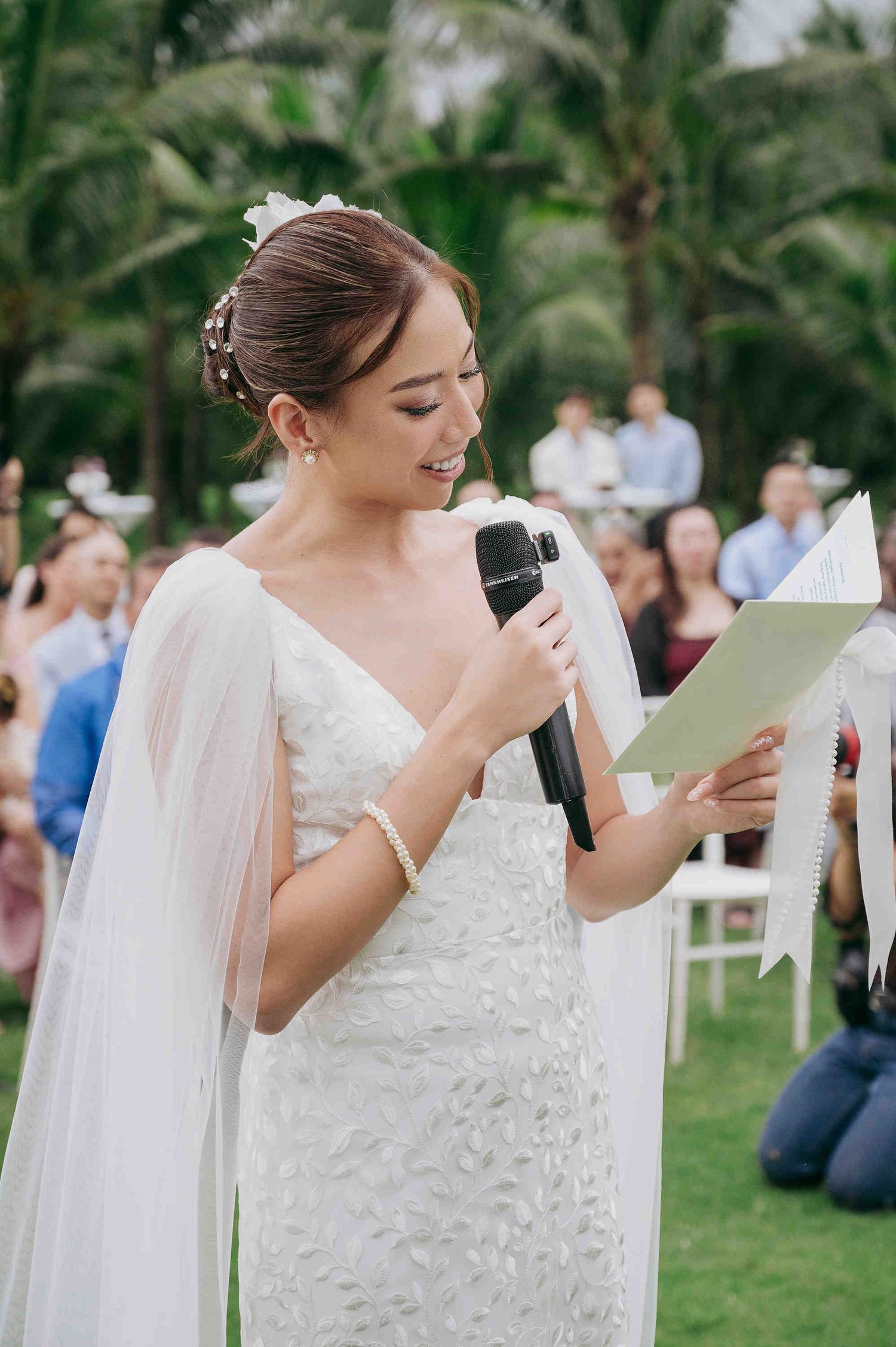Bride in white gown reads vows aloud at outdoor wedding ceremony, holding microphone and paper.