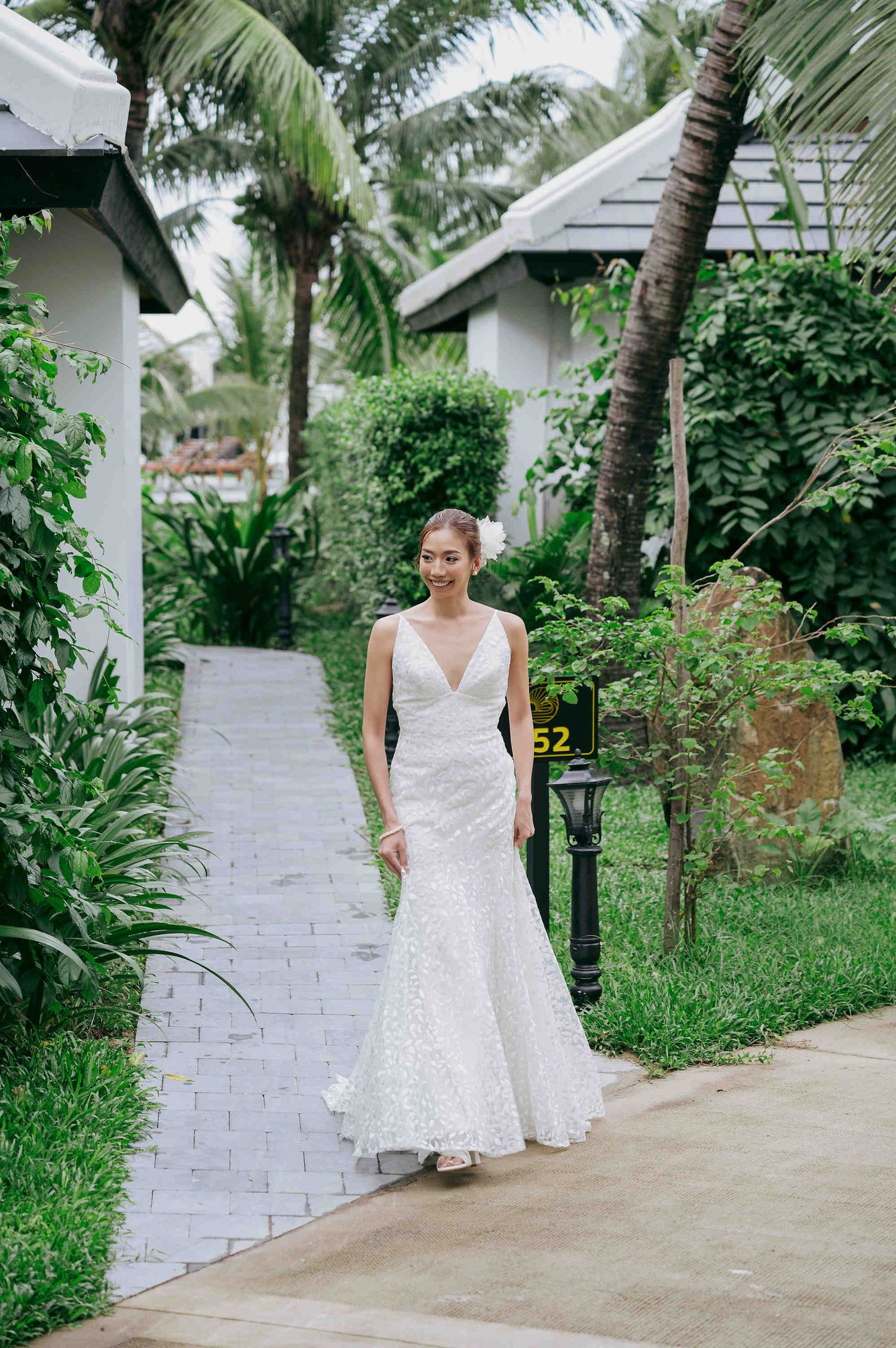 Bride in white lace mermaid gown walks along tropical garden pathway surrounded by palm trees and lush greenery.