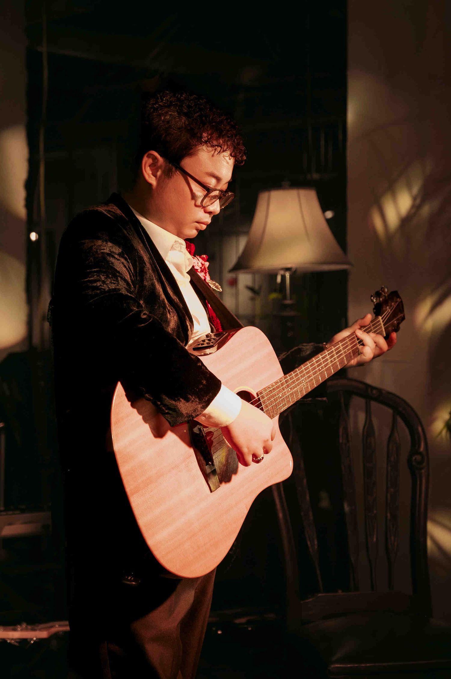 Young musician in a black blazer and bow tie playing an acoustic guitar in a warmly lit room.