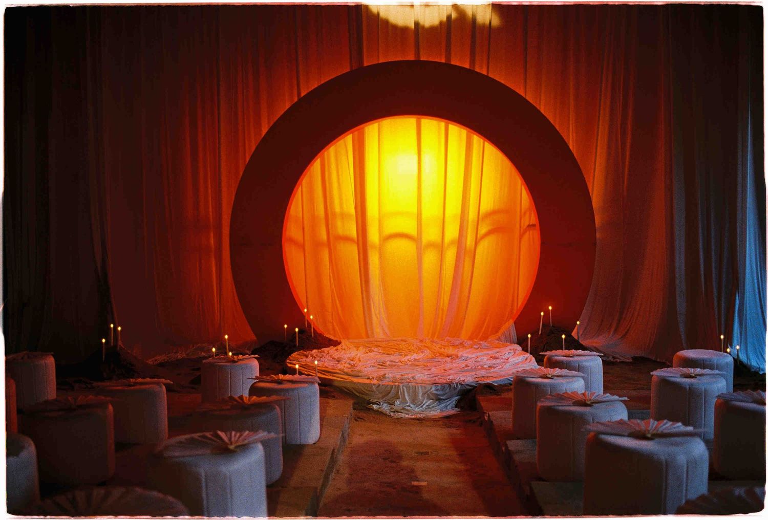Elegant event venue with glowing circular arch, draped orange curtains, candles, and white seating arrangements.