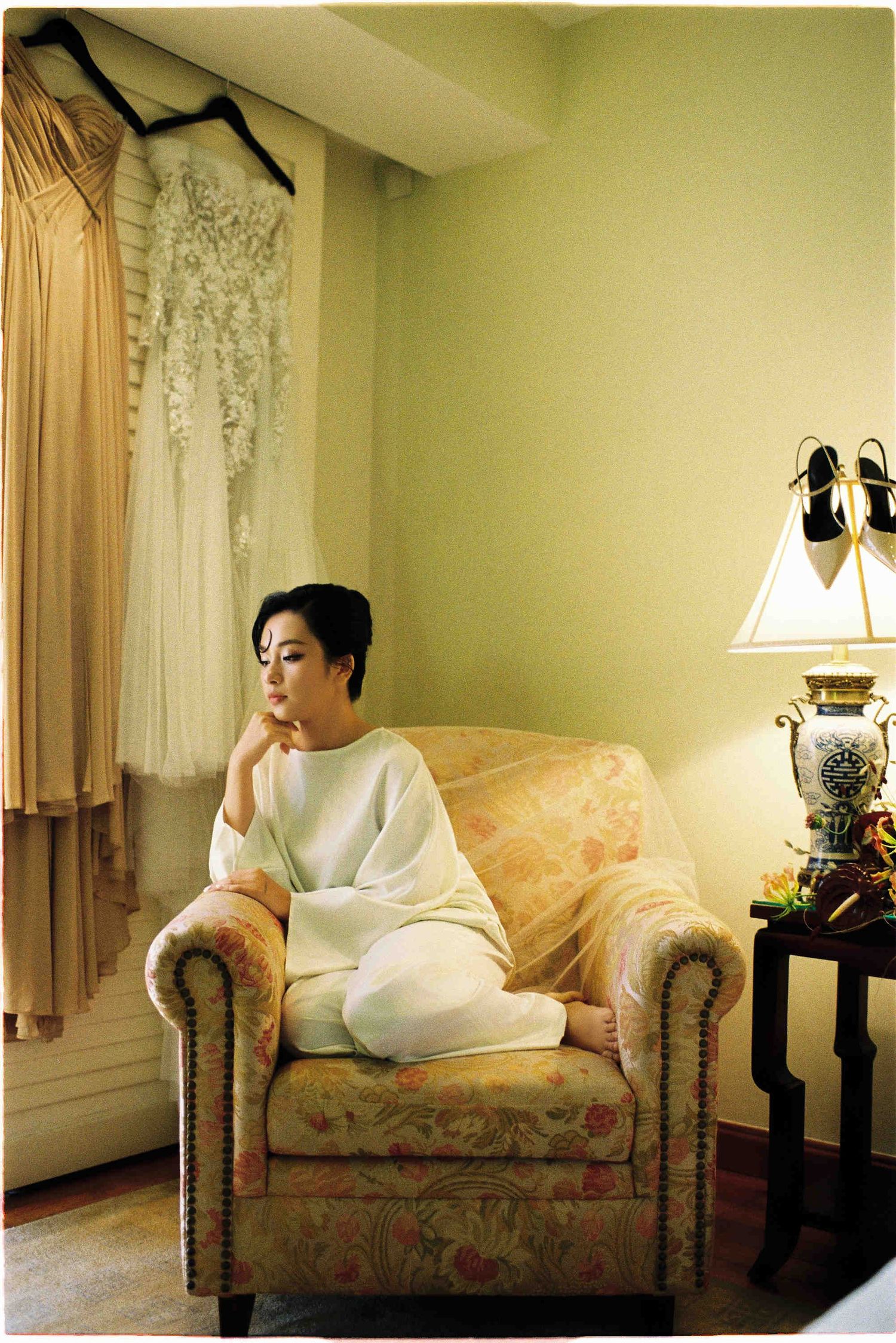 A woman in white sits cross-legged on a floral armchair beside a lamp, with dresses hanging by a curtained window.
