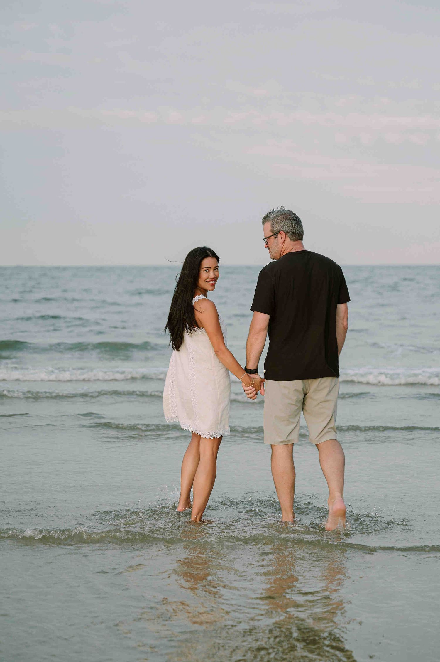 Da Nang family: couple wades together in shallow ocean waves at sunset, walking along the shoreline in a romantic moment, by AnhVuWedding.