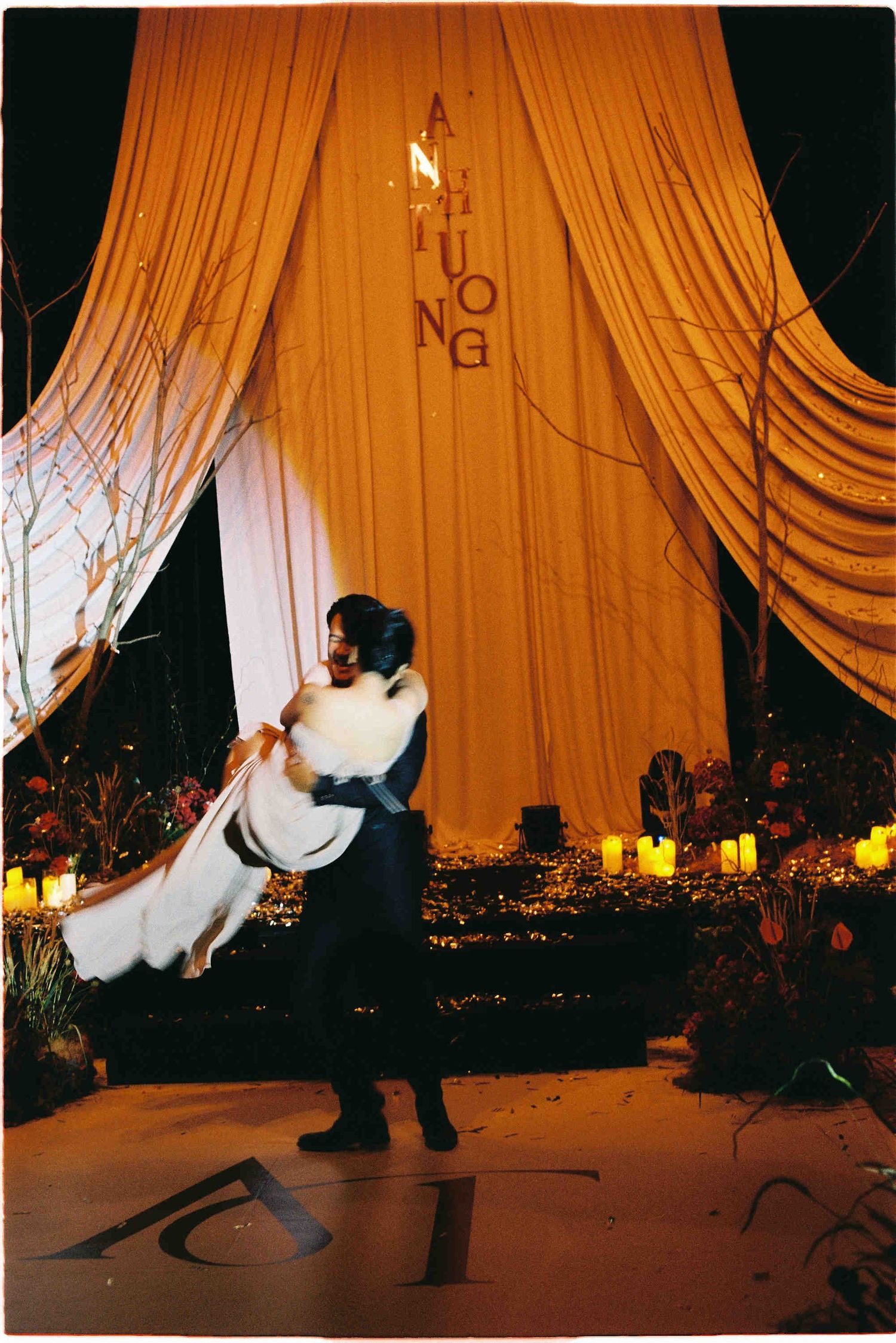 A groom lifts his bride in a white dress before a golden draped backdrop with candles at a romantic wedding reception.