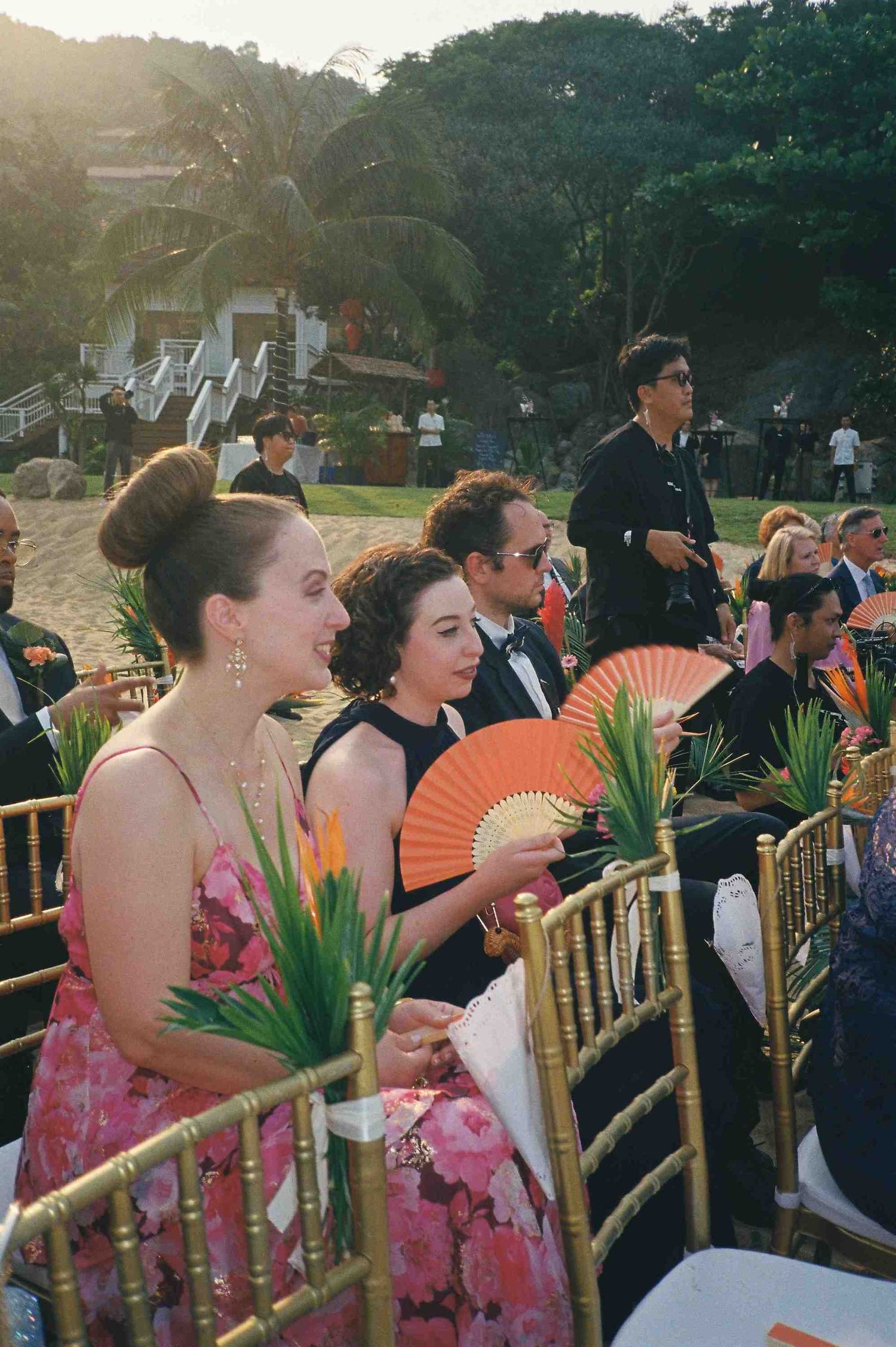 Lang Co wedding ceremony: wedding guests seated in bamboo chairs with orange paper fans at outdoor ceremony, by AnhVuWedding.