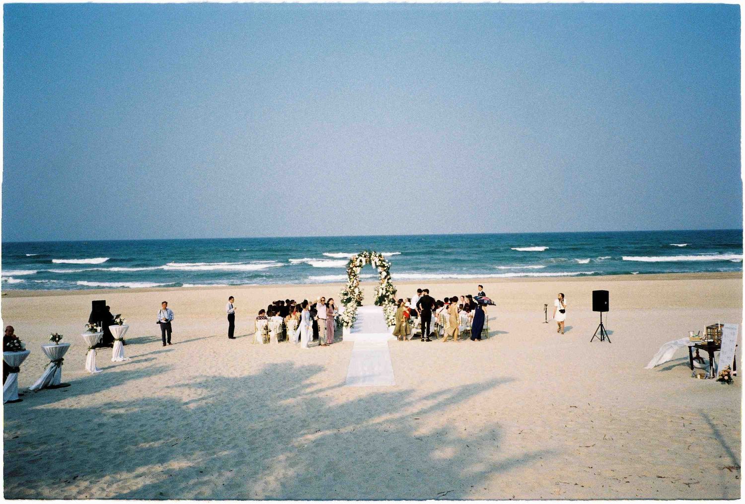 Outdoor beach wedding ceremony on sandy shore with ocean waves, floral arch, guests, and bridal party under clear blue sky.