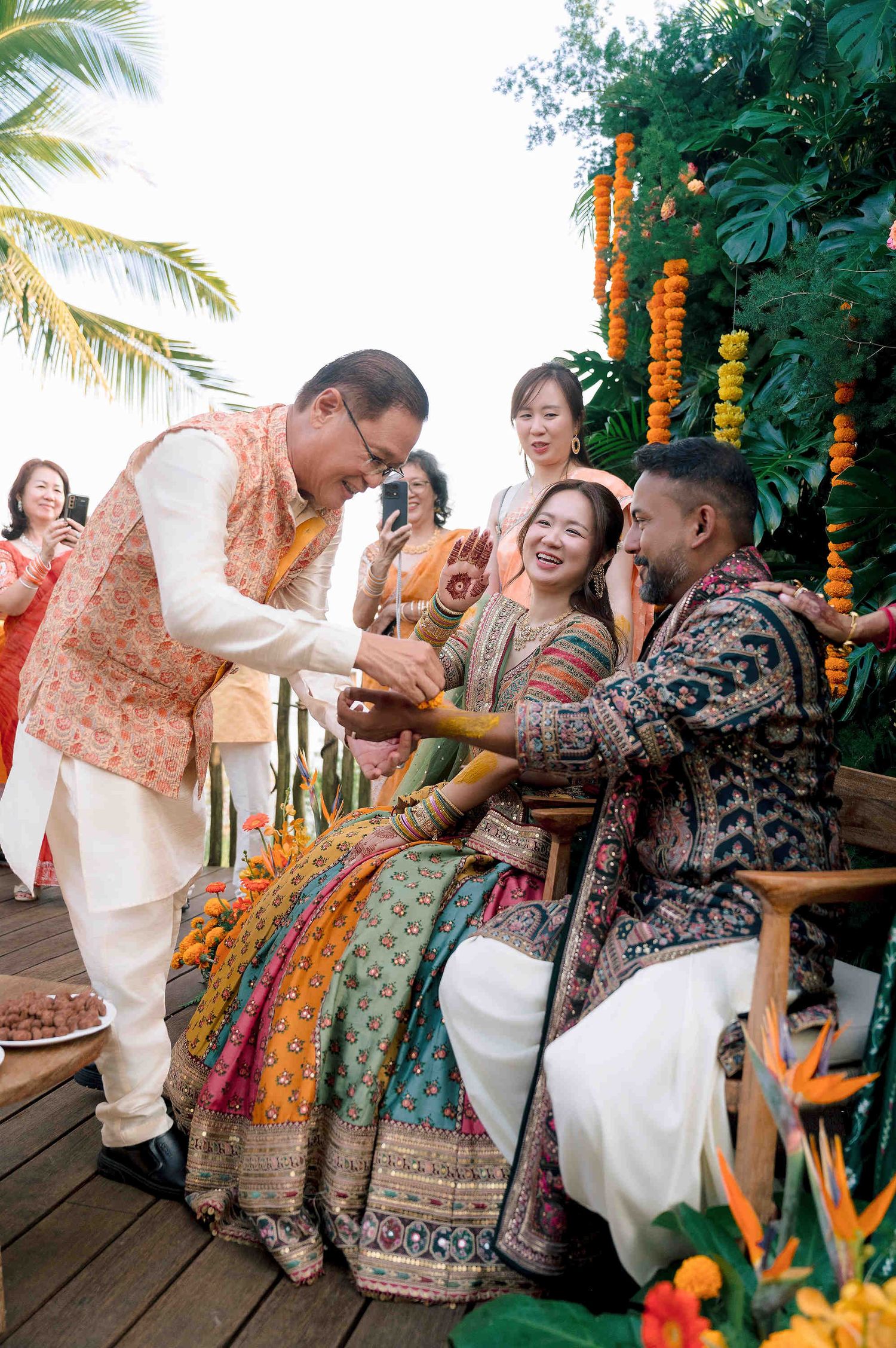 Indian wedding ceremony with guests in colorful traditional attire exchanging rings outdoors with tropical floral decor.