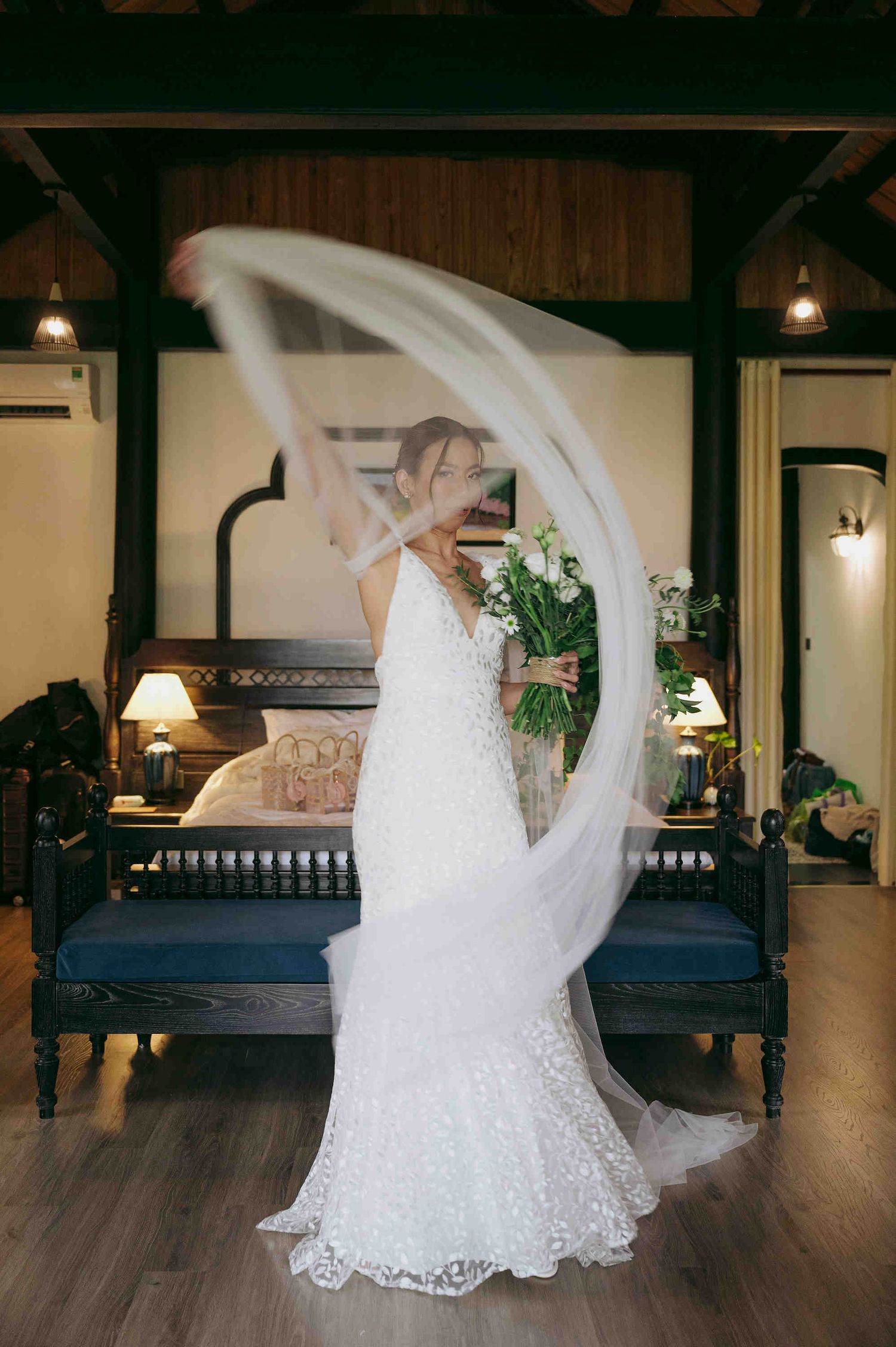 Bride in lace wedding gown twirling veil while holding bouquet in elegant dark-wood bedroom suite.