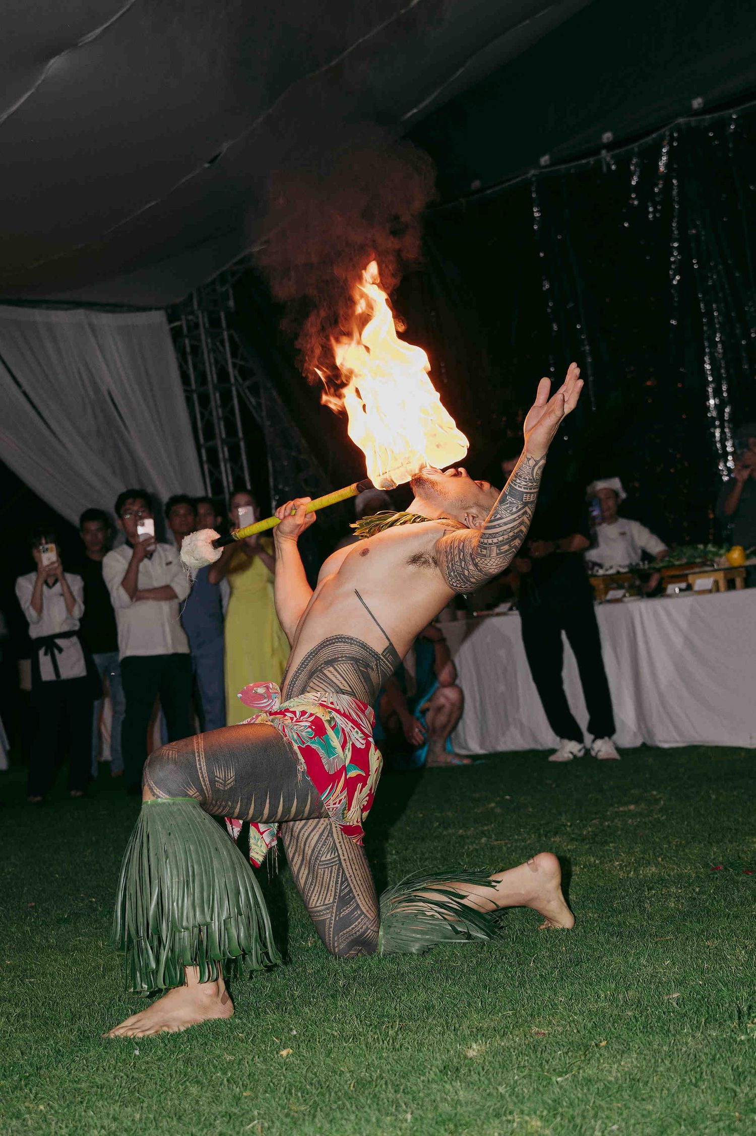 Tattooed fire dancer performs dramatic backbend while breathing fire at an outdoor evening event with spectators watching.