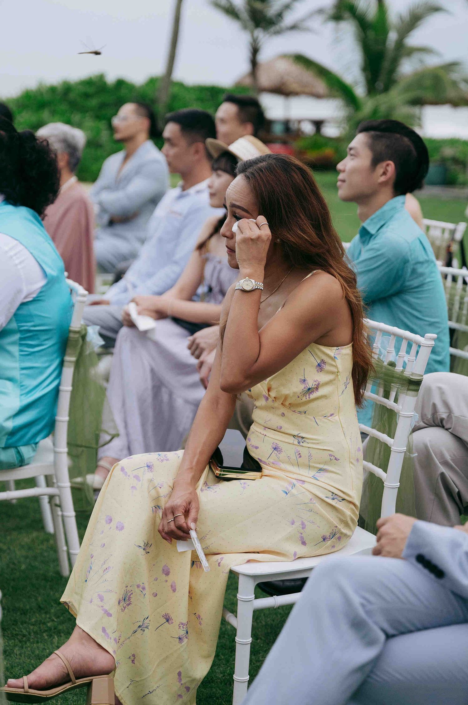 Emotional wedding guest in yellow floral dress wiping tears during outdoor ceremony, seated among other guests.