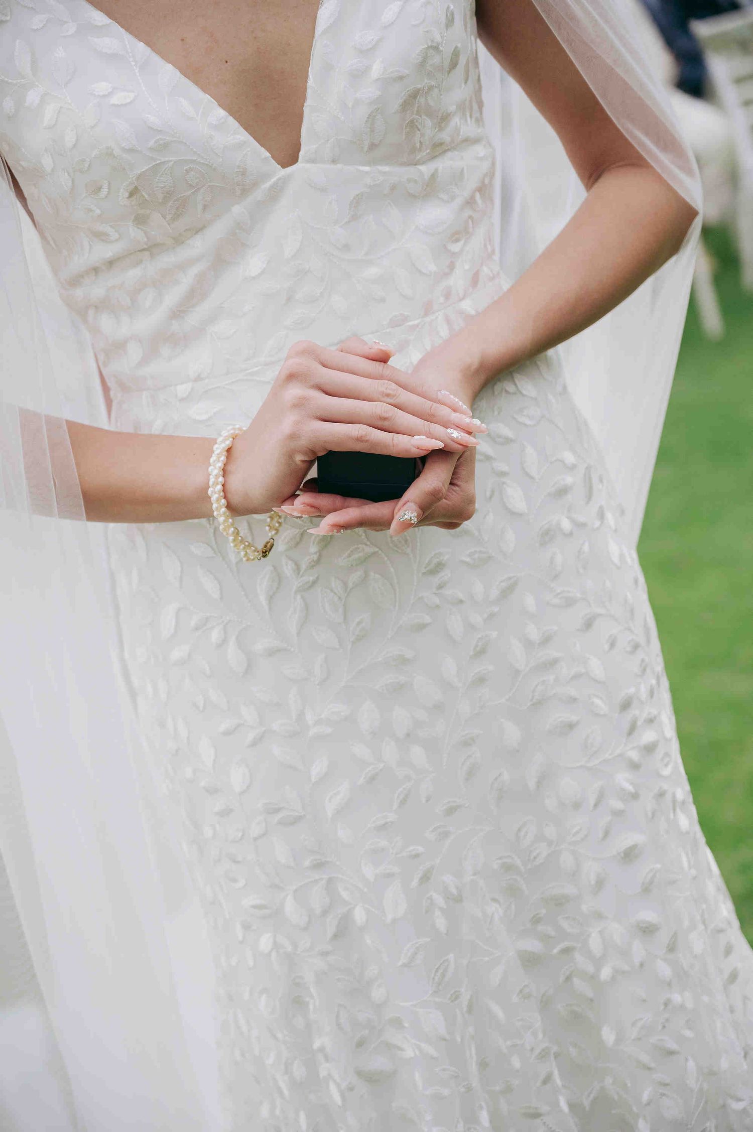 Bride in white lace wedding gown holding a small black ring box, wearing a pearl bracelet outdoors.