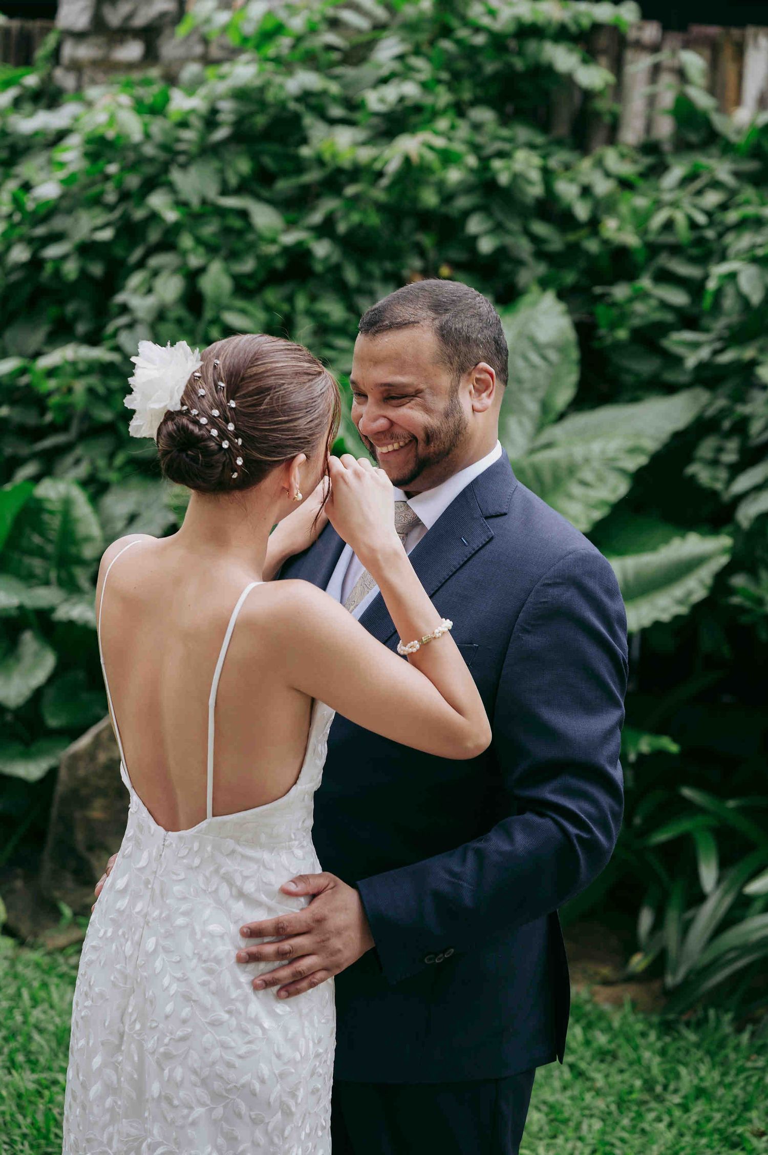 Groom in navy suit smiles warmly as bride in backless lace gown touches his face in lush garden.