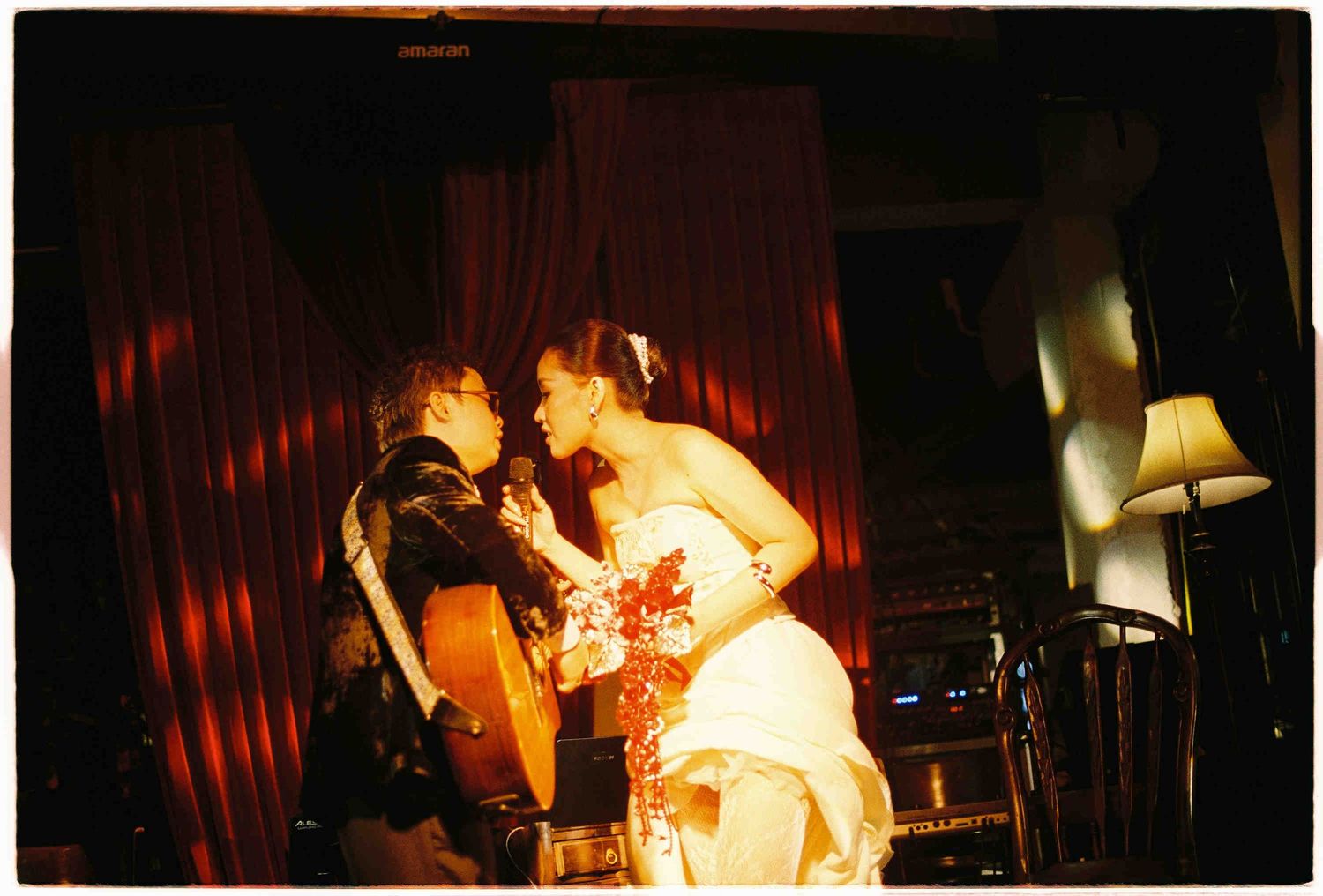Bride and groom sharing a romantic kiss at their wedding reception in a dimly lit venue with red curtains.