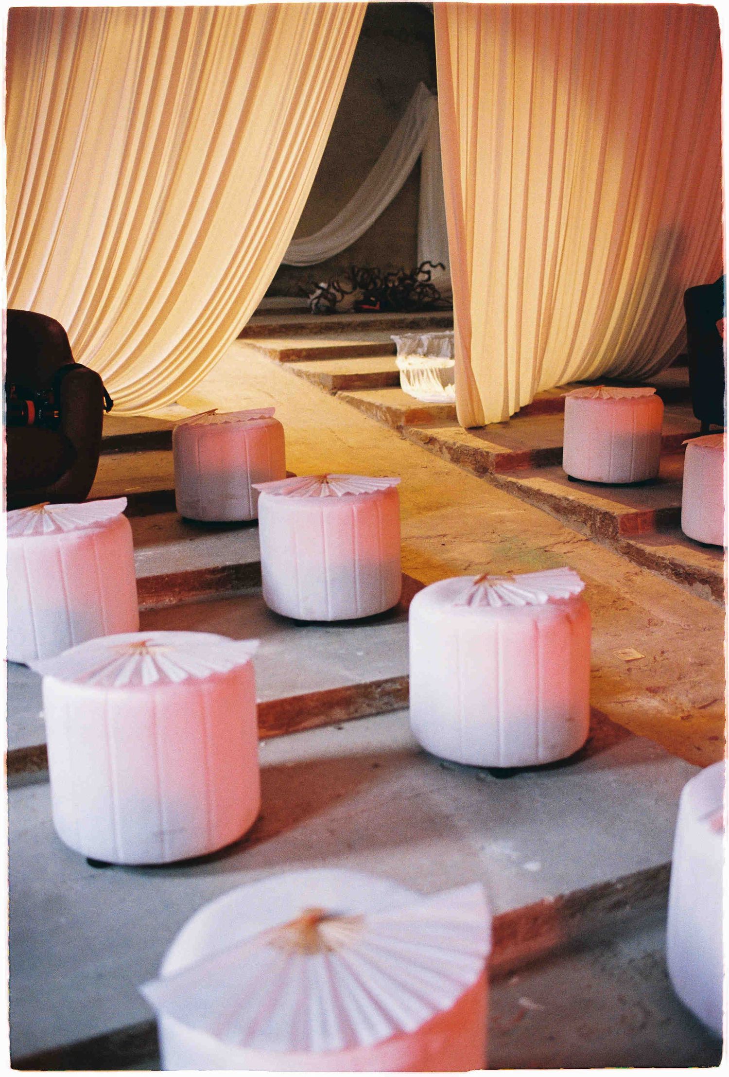 Elegant event setup with white draped ottomans on stone floor beneath flowing cream curtains.