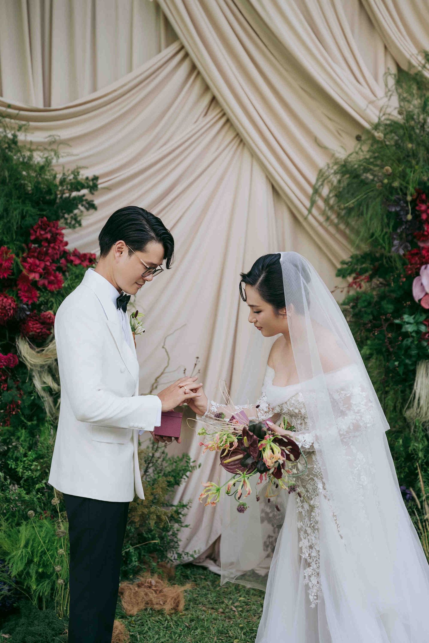 Couple exchanging rings at elegant outdoor wedding ceremony with floral arch backdrop.
