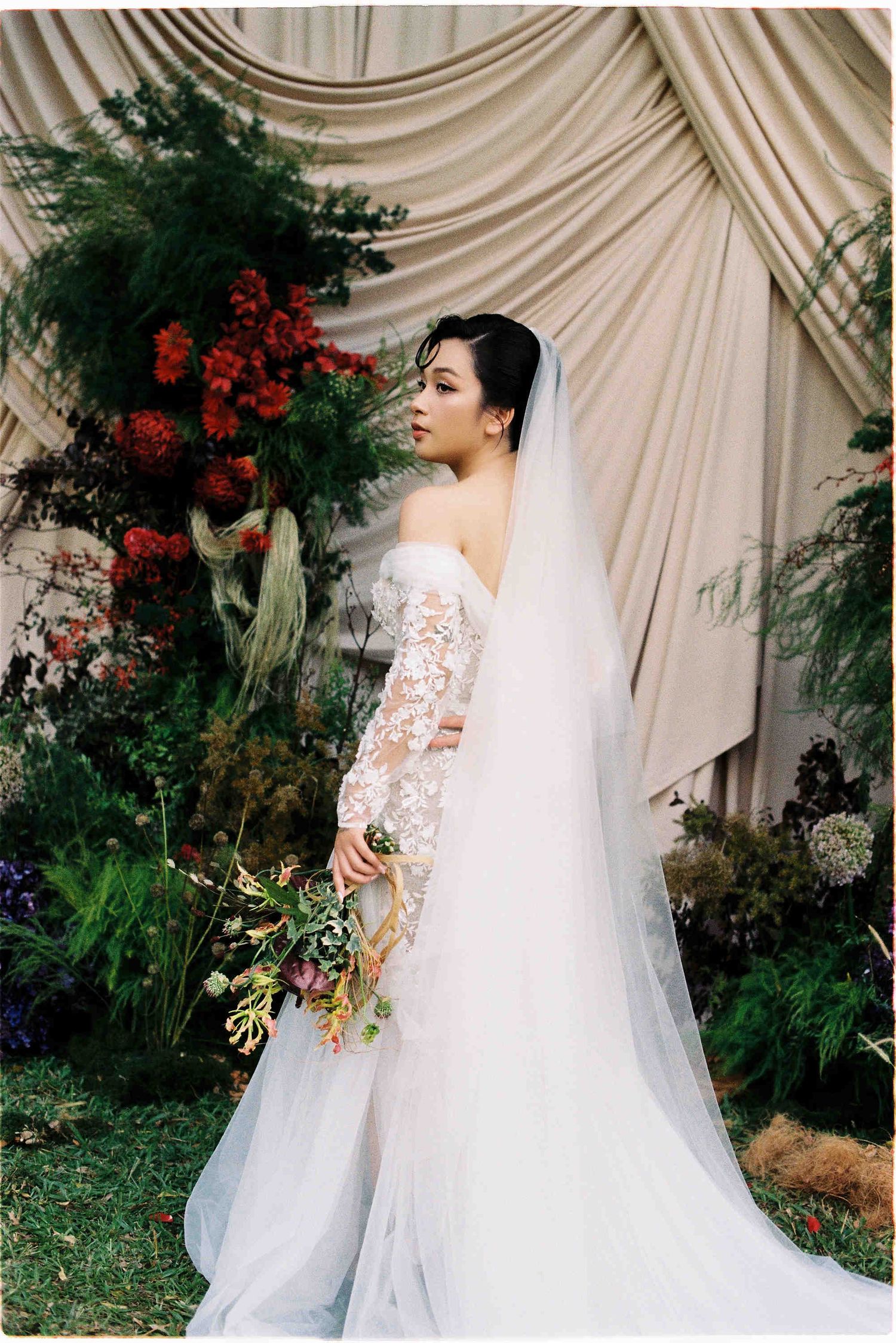 Bride in off-shoulder lace wedding gown with long veil holds bouquet before floral backdrop.