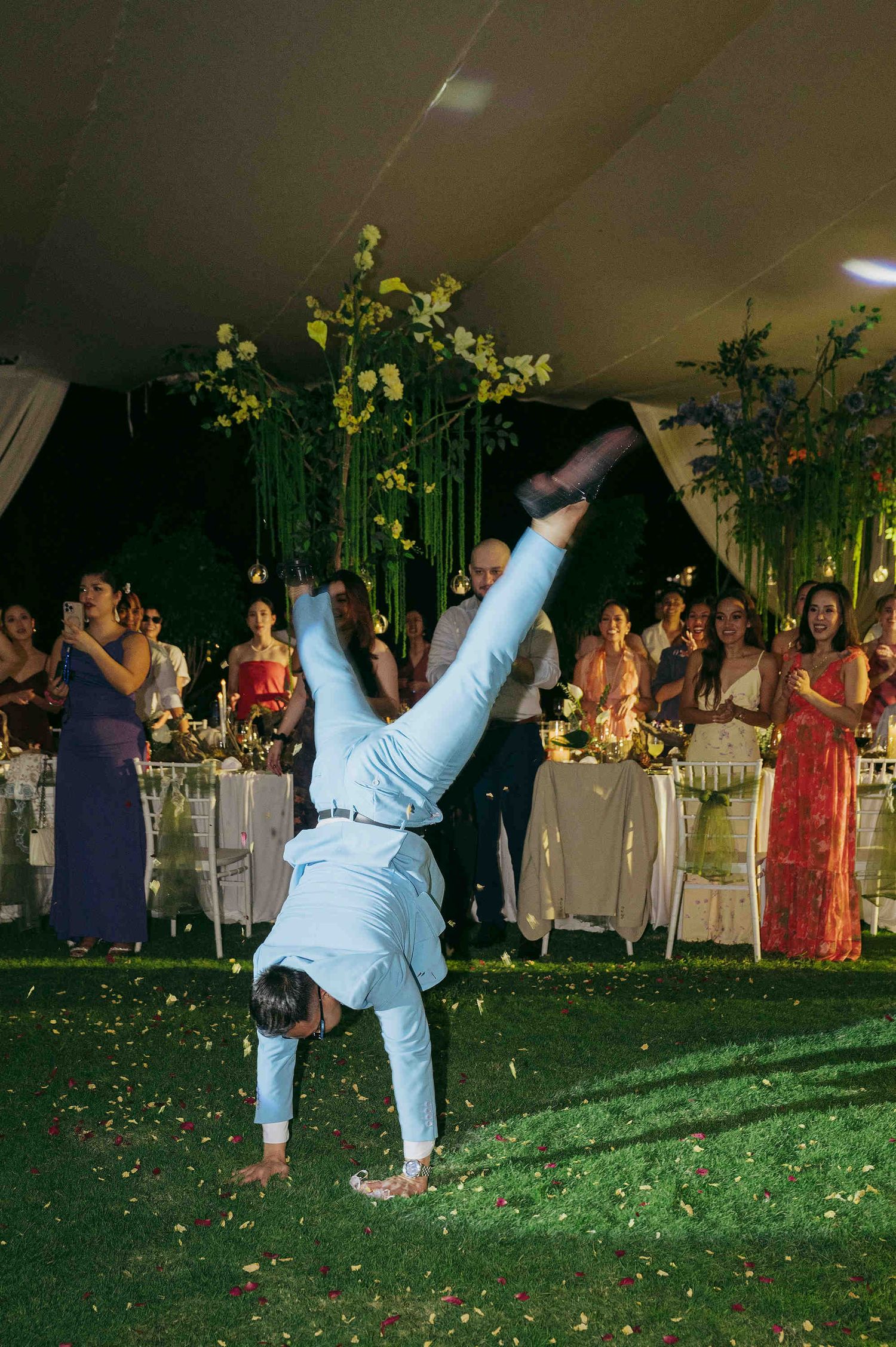 A guest performs a handstand at an elegant outdoor wedding reception under a floral-decorated tent.