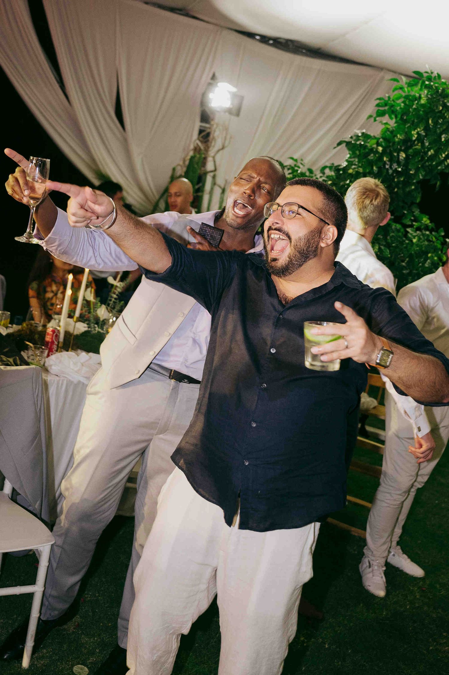 Two men celebrating at an outdoor wedding reception, dancing and holding drinks under a draped tent at night.
