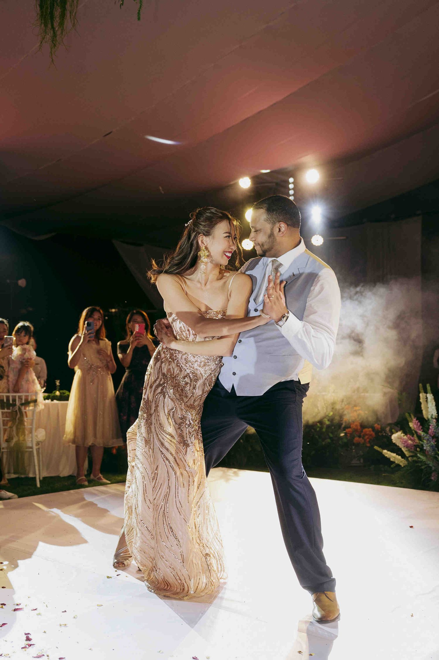Couple sharing their first dance at a wedding reception, bride in gold sequin gown, groom in white shirt and navy pants.