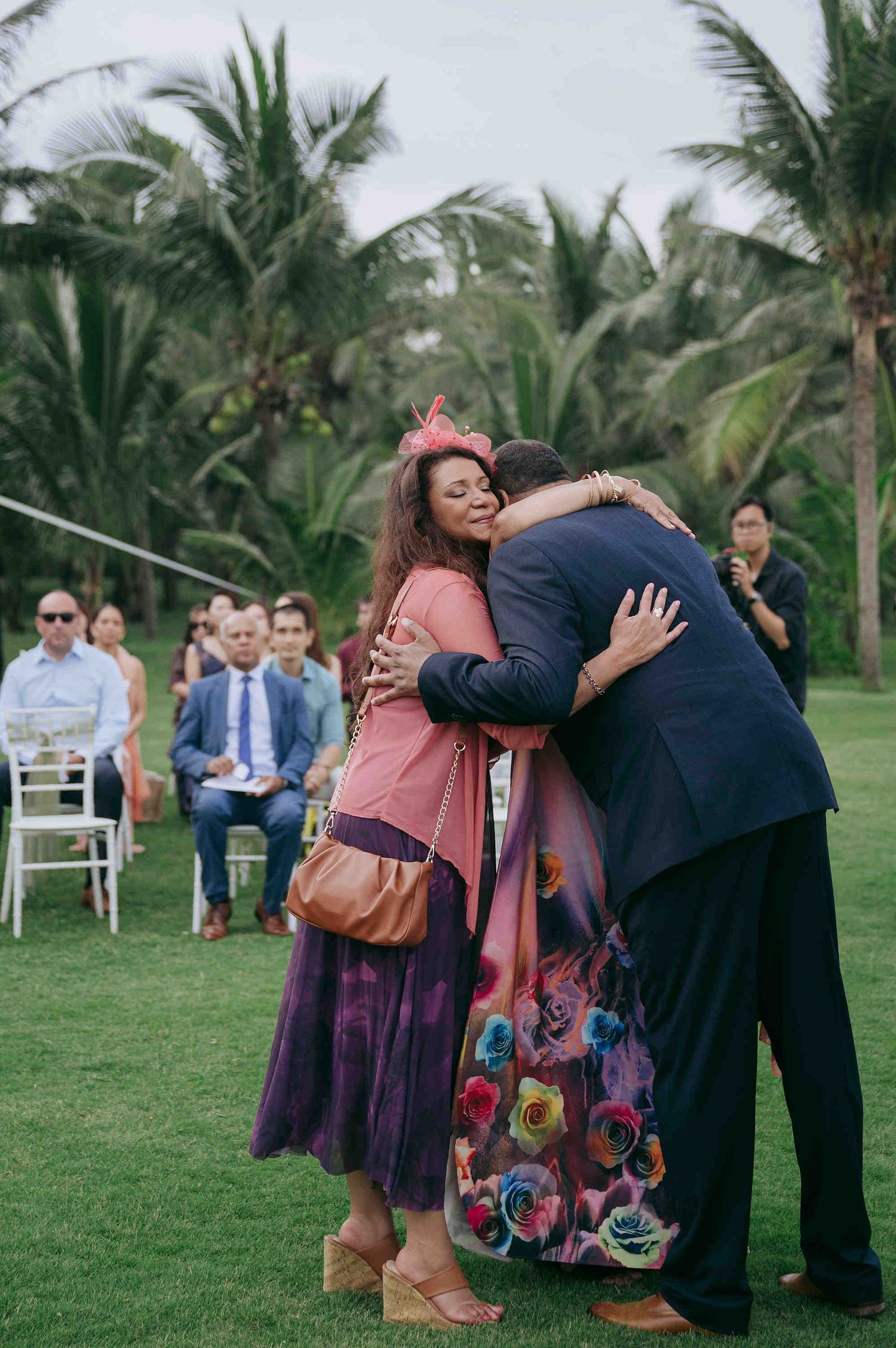 A woman in a floral dress hugs a man in a navy suit at an outdoor wedding ceremony with palm trees.