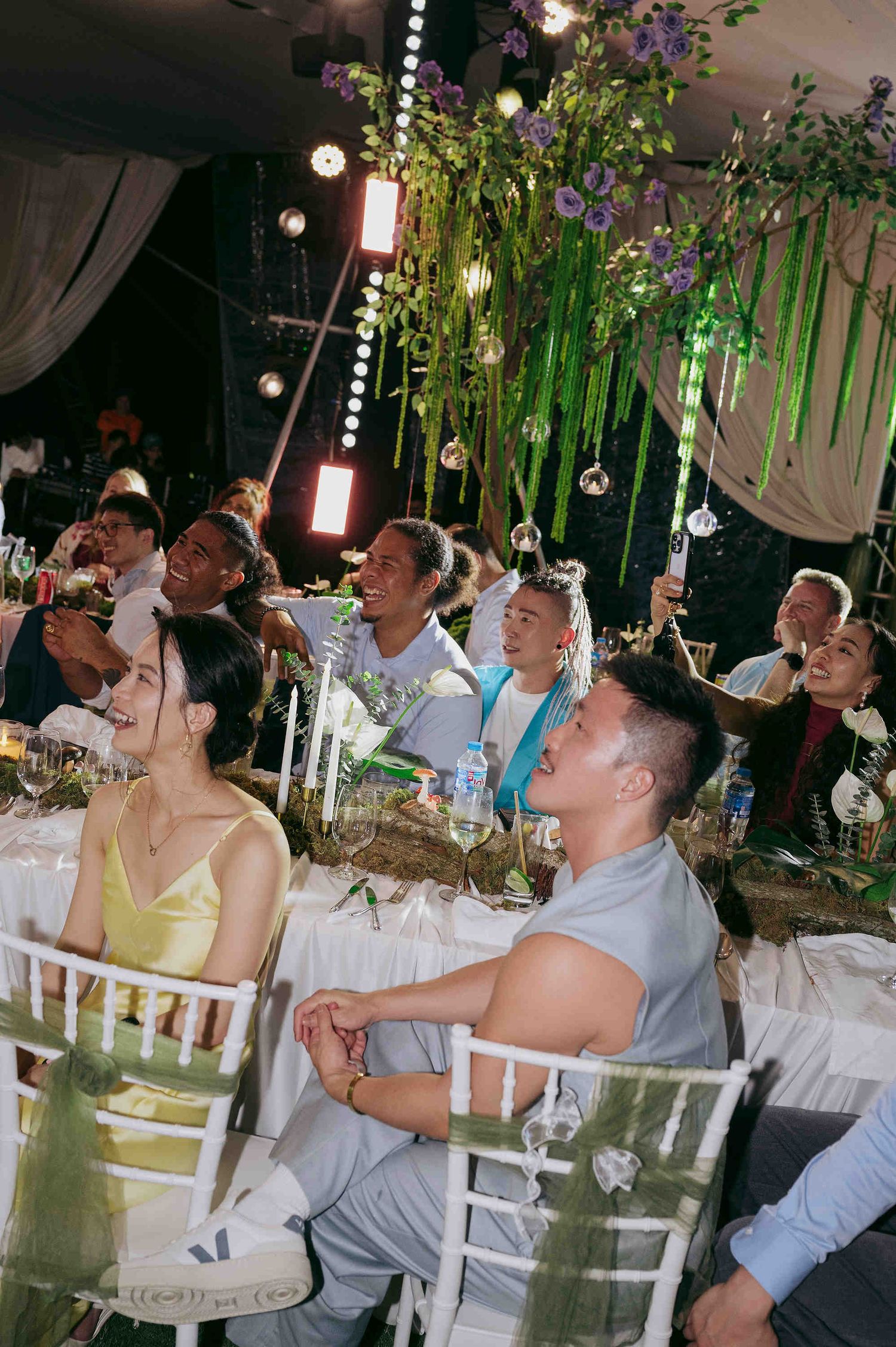 Guests seated at elegantly decorated wedding reception tables, looking up at lush floral ceiling installations.