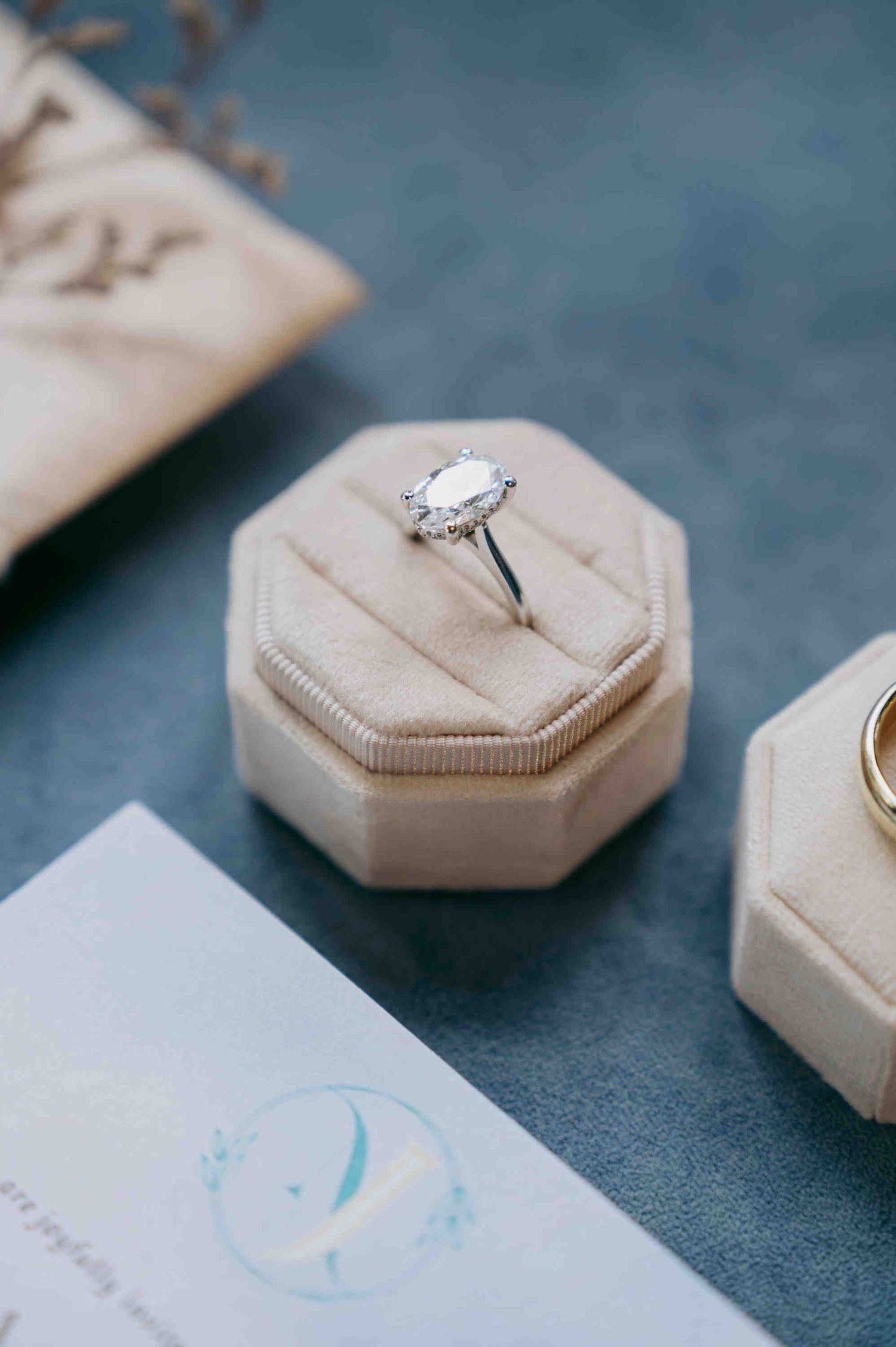 Elegant oval diamond solitaire engagement ring displayed in a beige velvet octagon ring box on a blue surface.