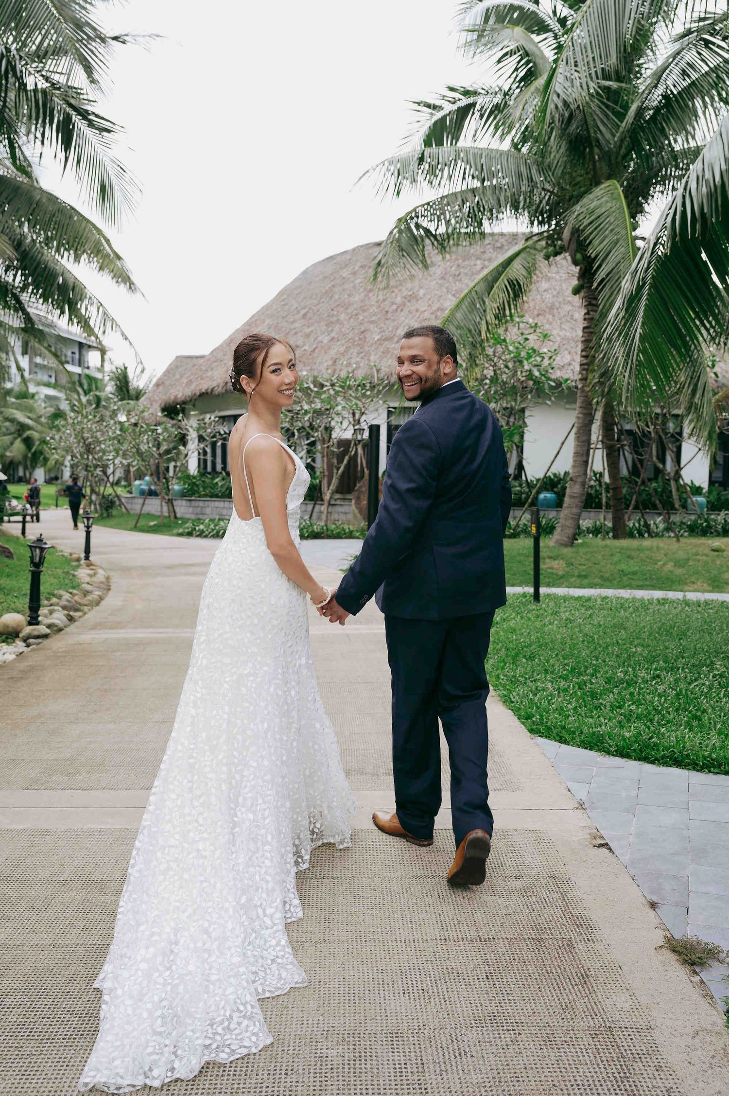 Newlyweds holding hands on tropical resort path, bride in backless lace gown smiling over shoulder.