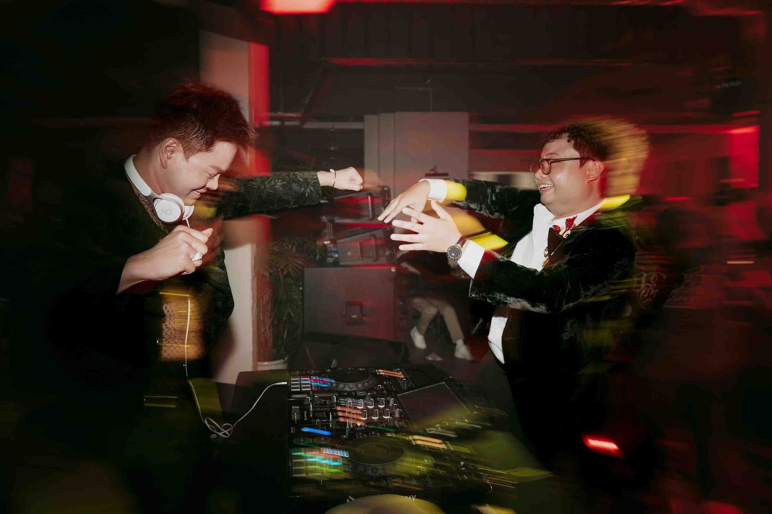 Two DJs in suits fist-bumping over DJ equipment at a dimly lit event with red ambient lighting.