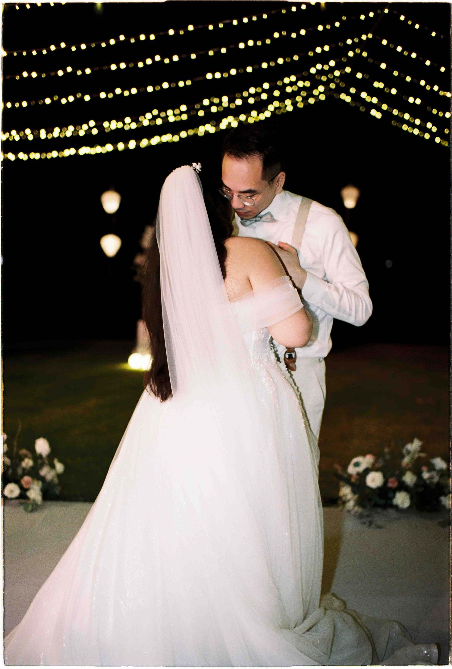 Bride and groom share first dance under twinkling string lights at elegant outdoor night wedding ceremony.
