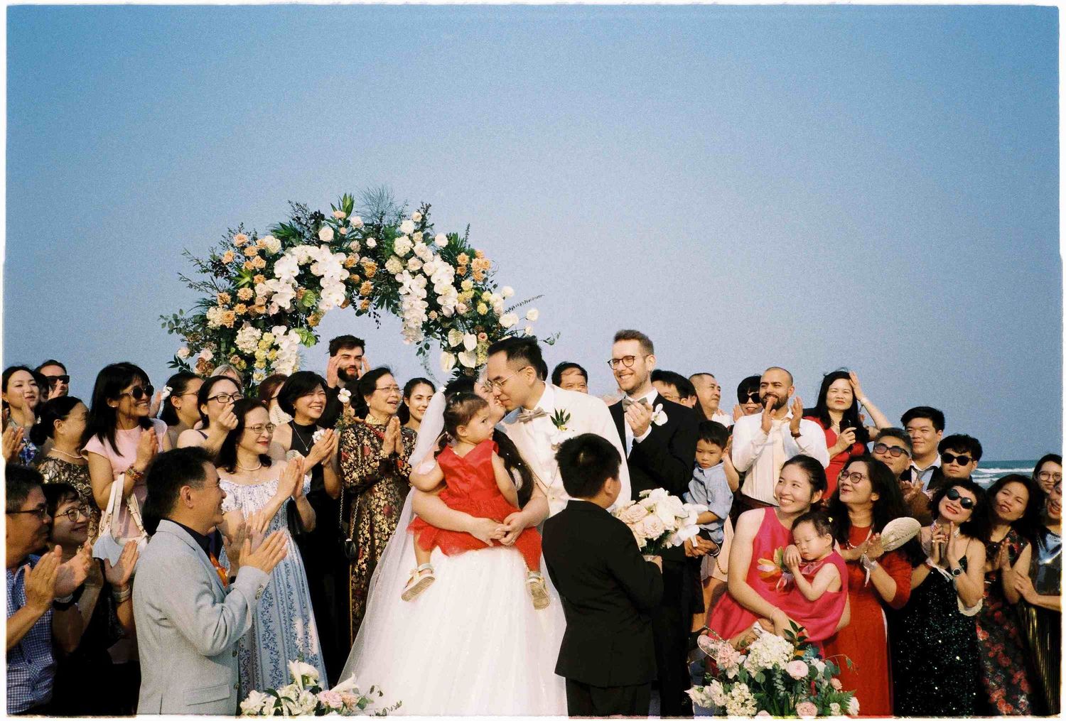 Joyful wedding ceremony with bride and groom kissing under floral arch, surrounded by cheering guests outdoors.