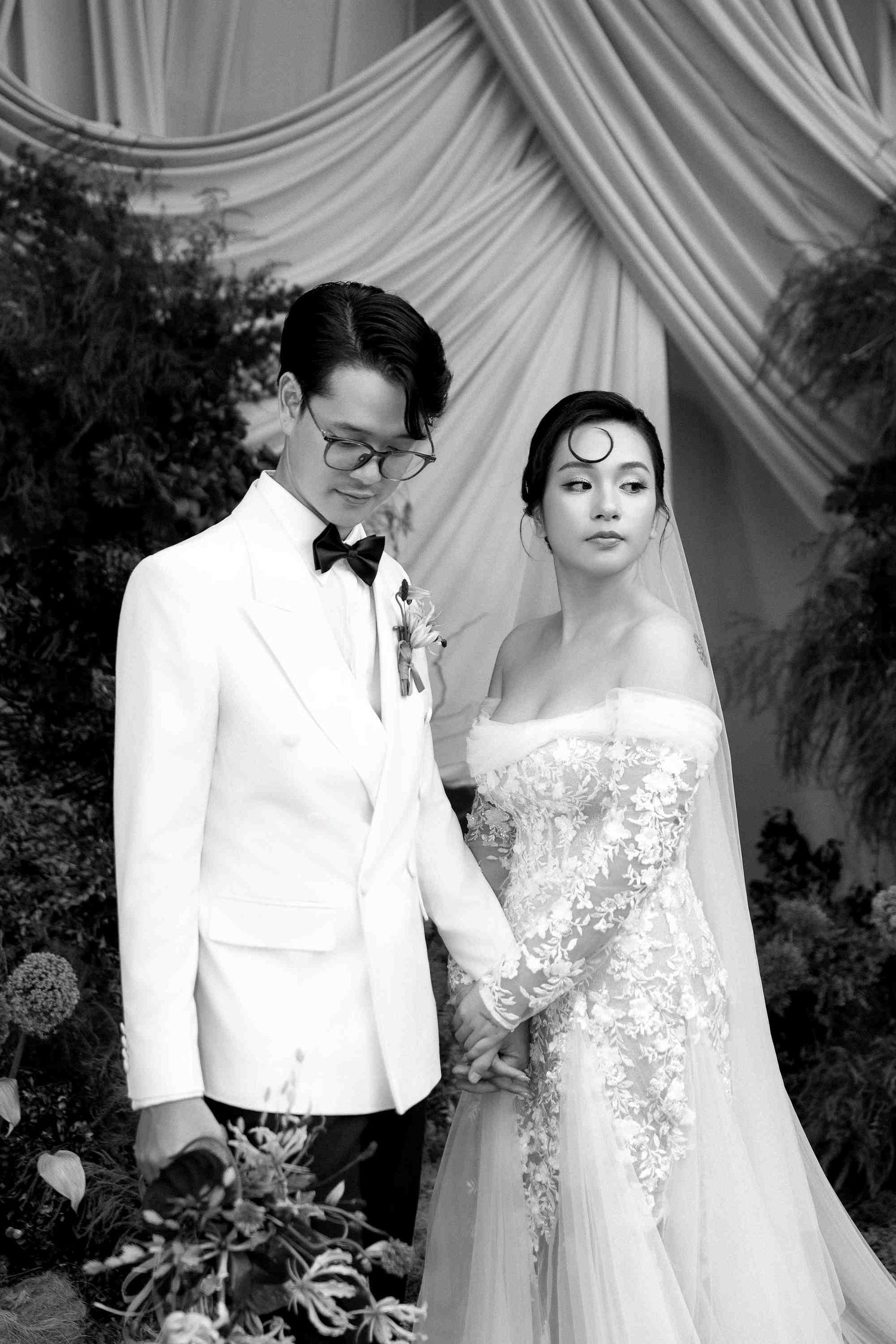 Black and white wedding photo of an elegant couple, groom in white tuxedo and bride in lace gown, holding hands.