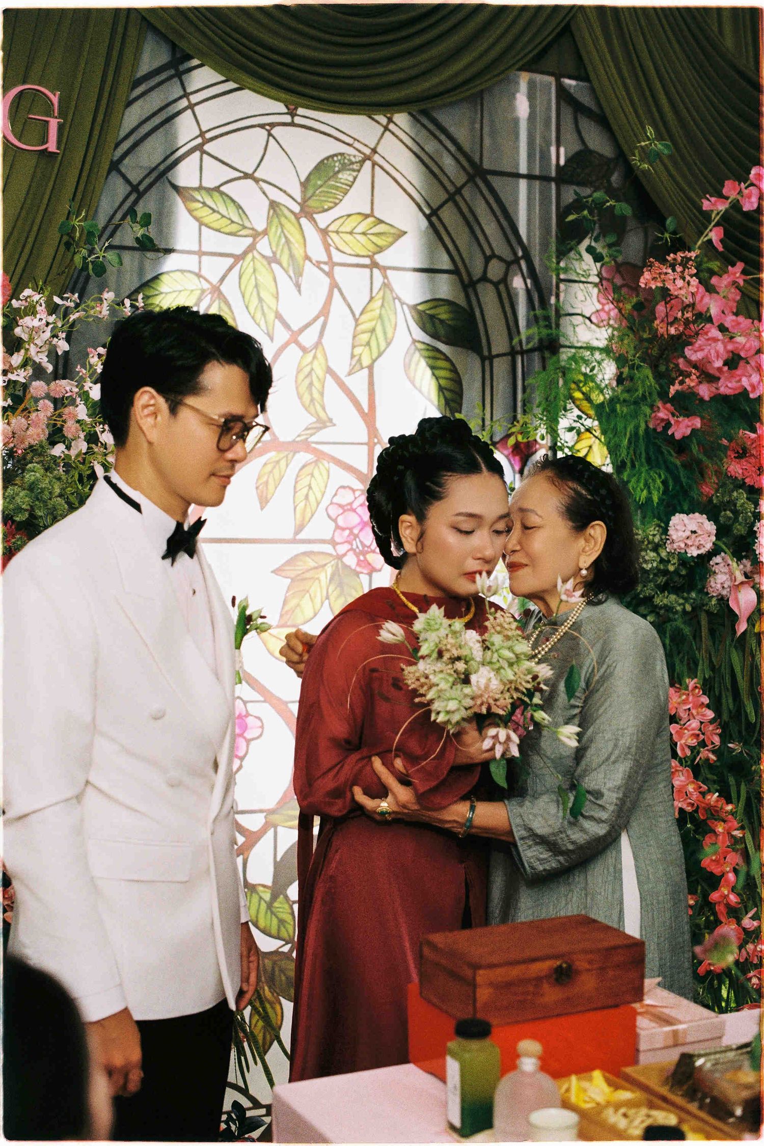 Emotional Vietnamese wedding ceremony with bride in red ao dai embracing elder woman while groom watches in white suit.