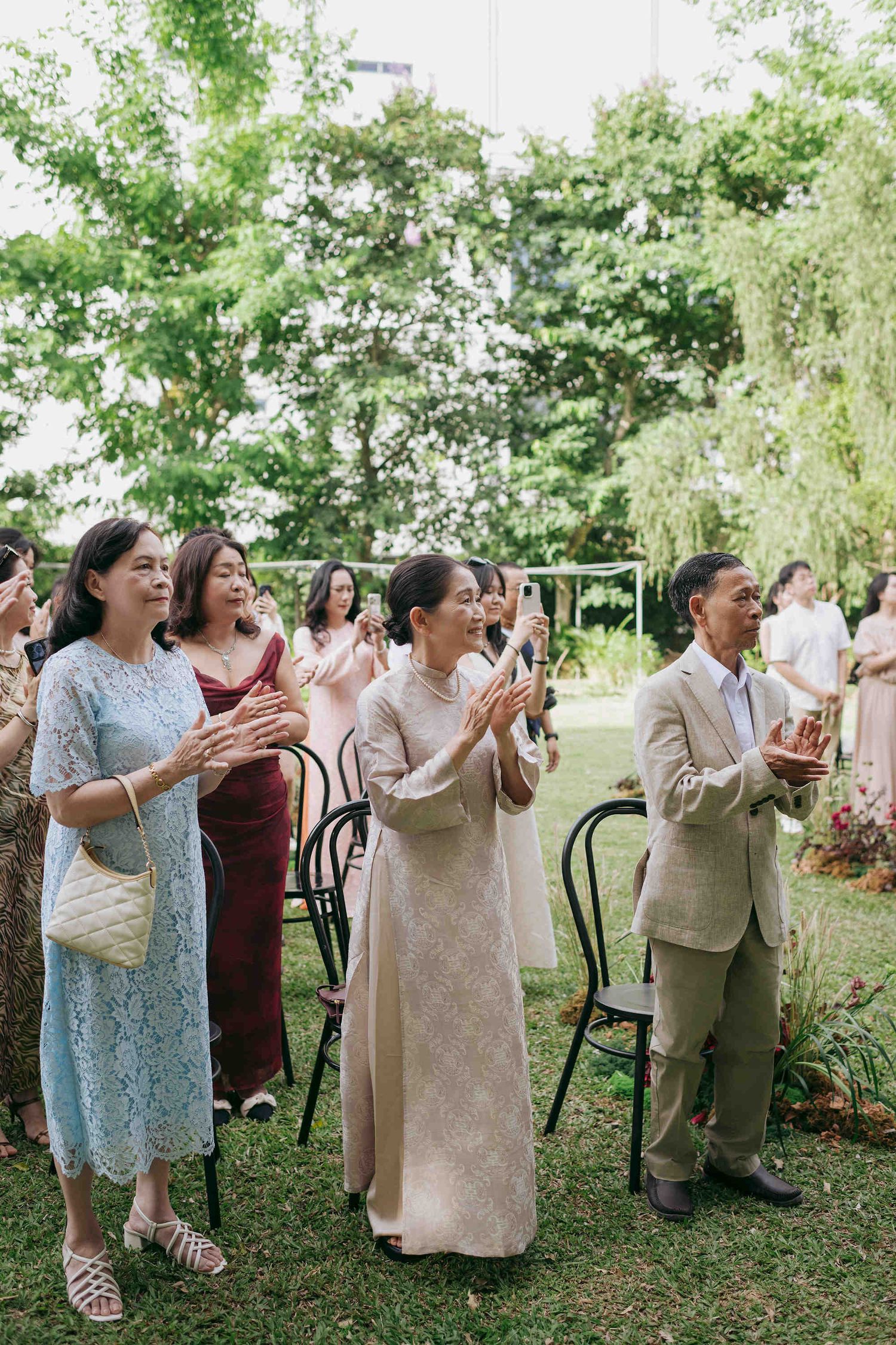 Wedding guests clapping outdoors at a garden ceremony, dressed in elegant attire amid lush green trees.