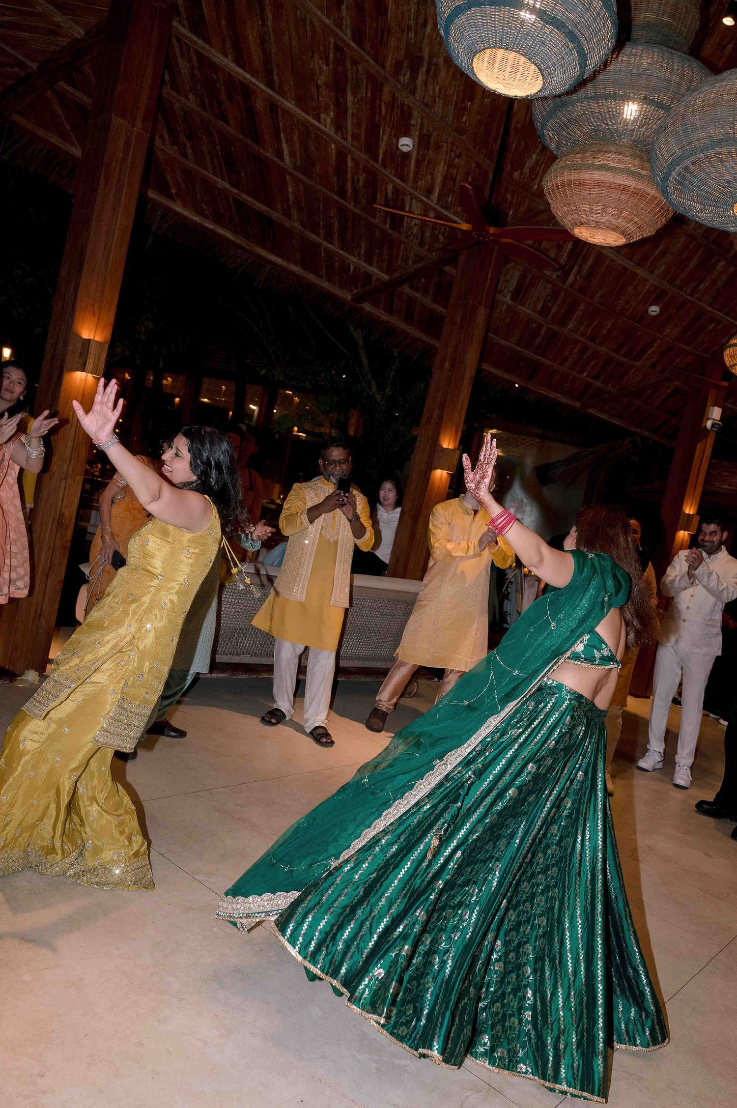 Women in vibrant green and gold lehengas dancing at an Indian wedding celebration under decorative hanging lanterns.