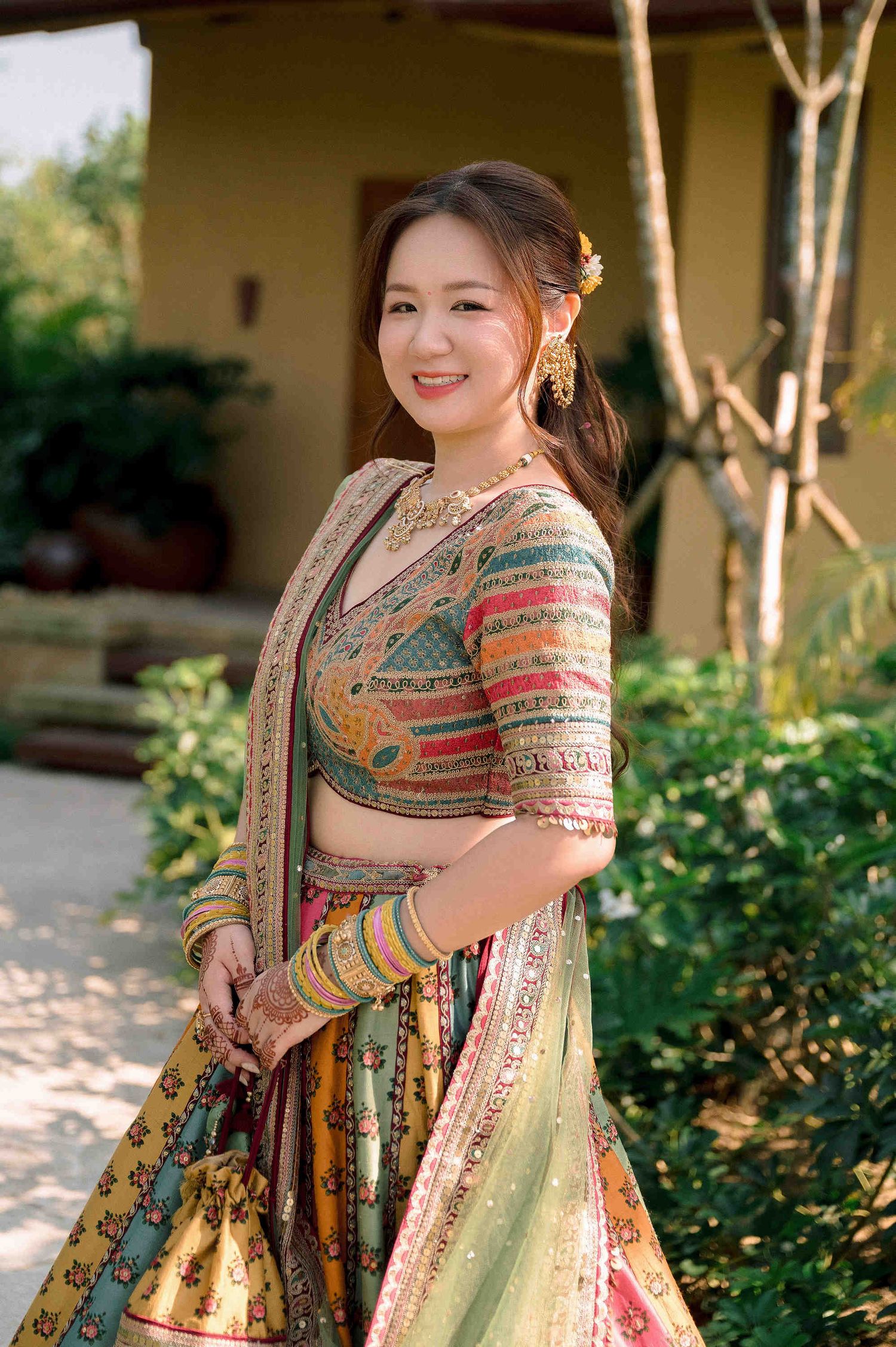 A smiling woman in a colorful traditional Indian lehenga choli with bangles and floral hair accessory, posing outdoors.