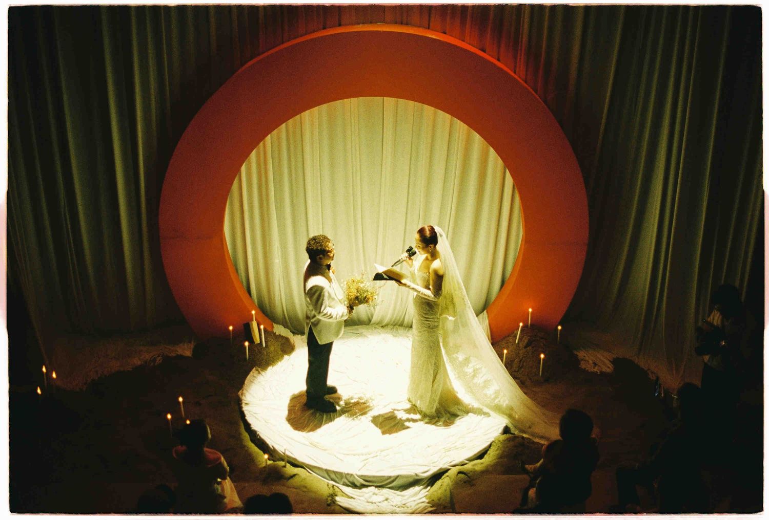 Couple exchanging vows on a circular stage with candles and a glowing arch backdrop in a dramatic wedding ceremony.