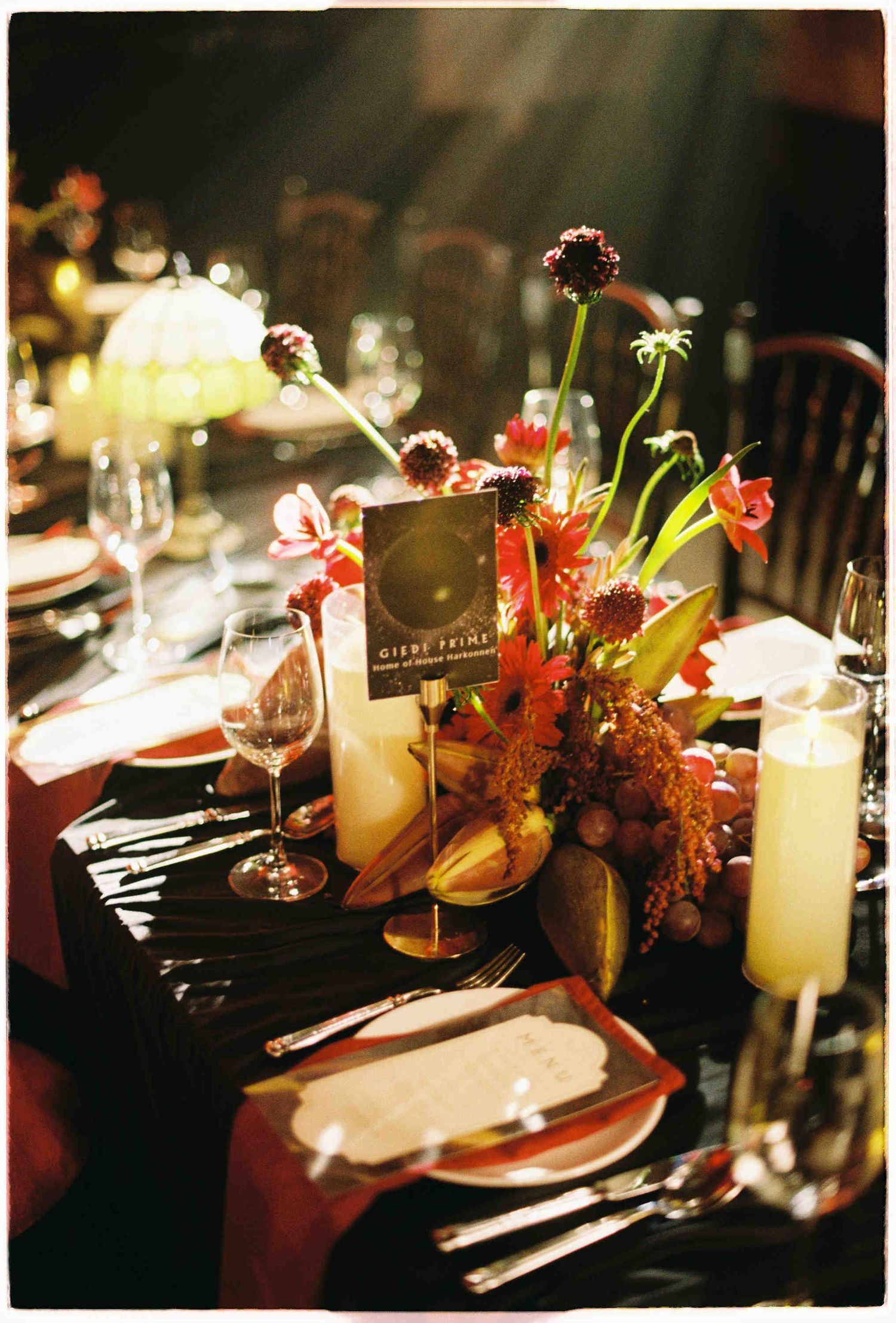 Romantic dinner table with tall candles, red floral arrangements, wine glasses, and place settings in warm ambient lighting.