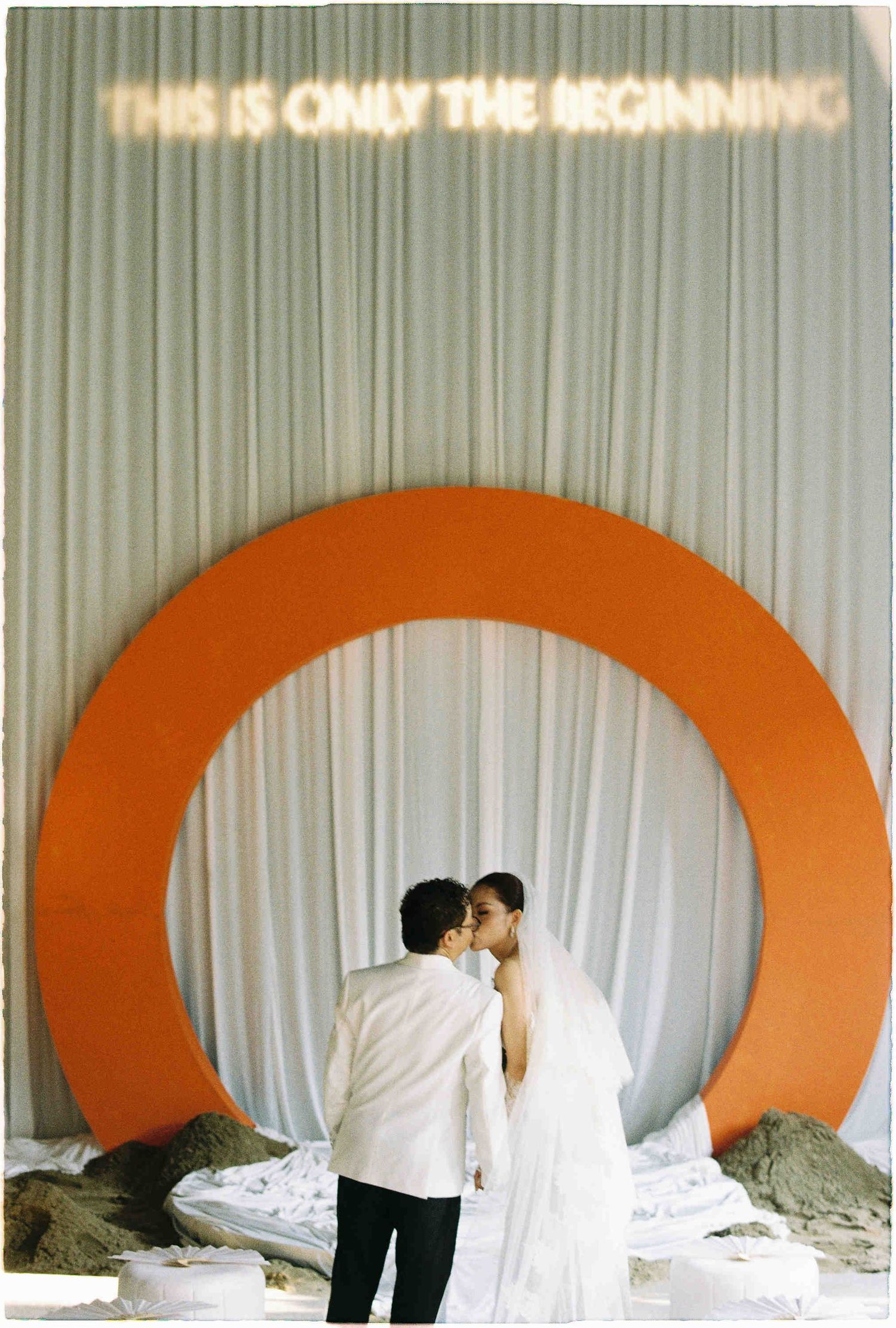Couple kissing under orange circular arch with neon sign reading This Is Only The Beginning at wedding.