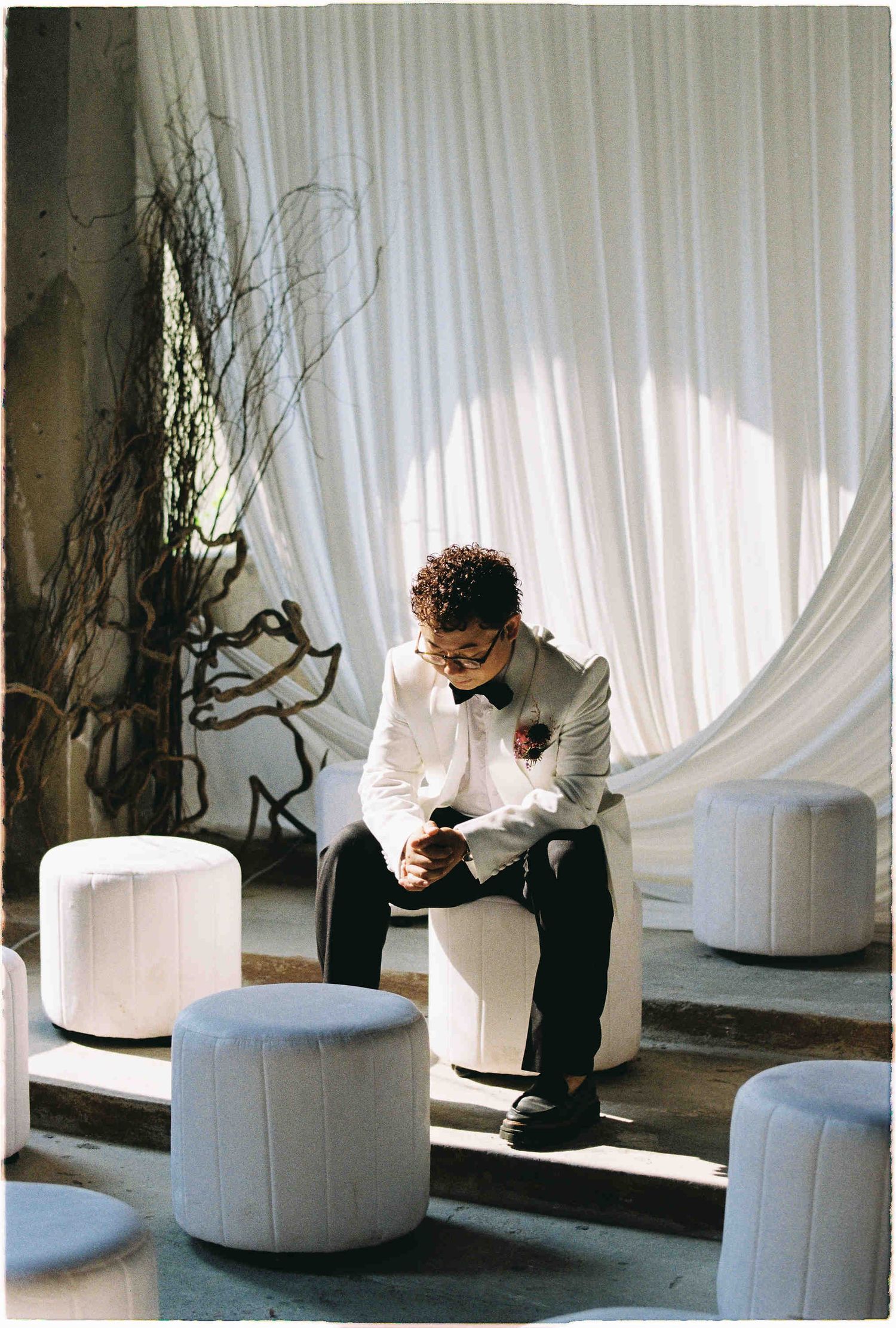 A groom in a white tuxedo jacket sits on a round ottoman, adjusting his cufflinks in an elegant draped venue.