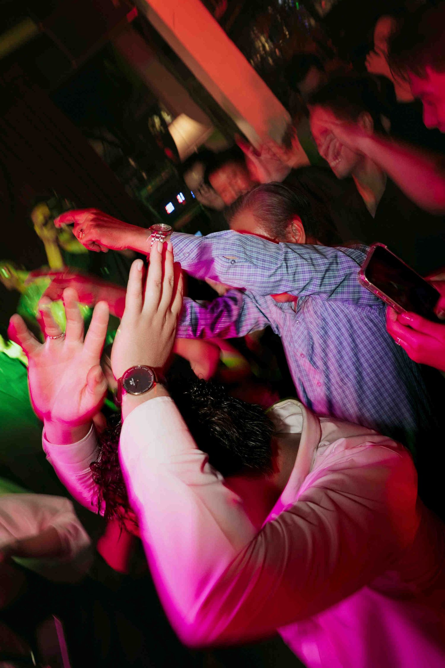 Crowd of people dancing energetically under colorful pink and red lights at a nightclub party.
