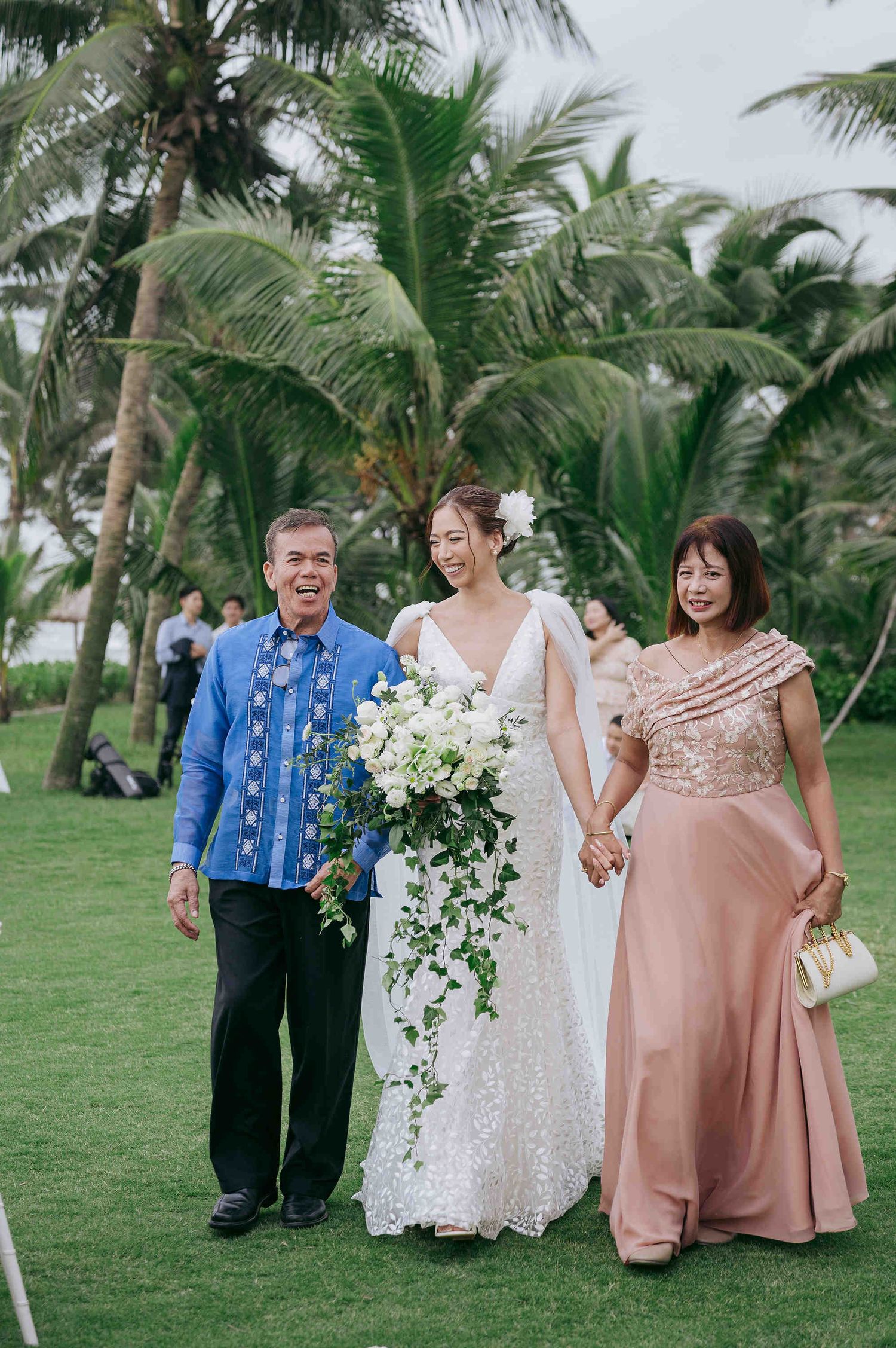 Bride in white gown with cascading bouquet walks with parents on tropical lawn at outdoor beach wedding.