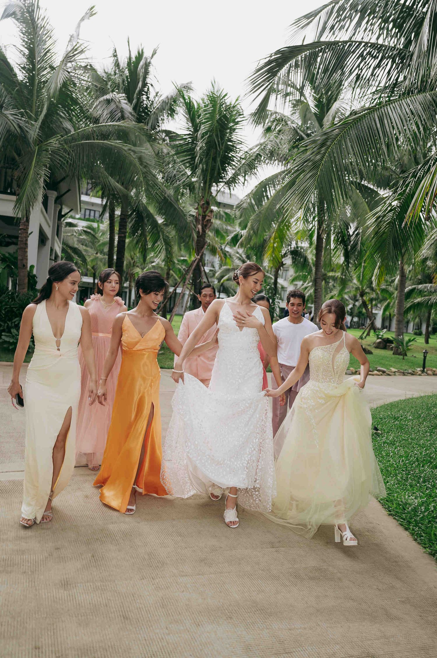 Bride in white lace gown walks with bridesmaids in pastel dresses along a tropical palm tree-lined path.