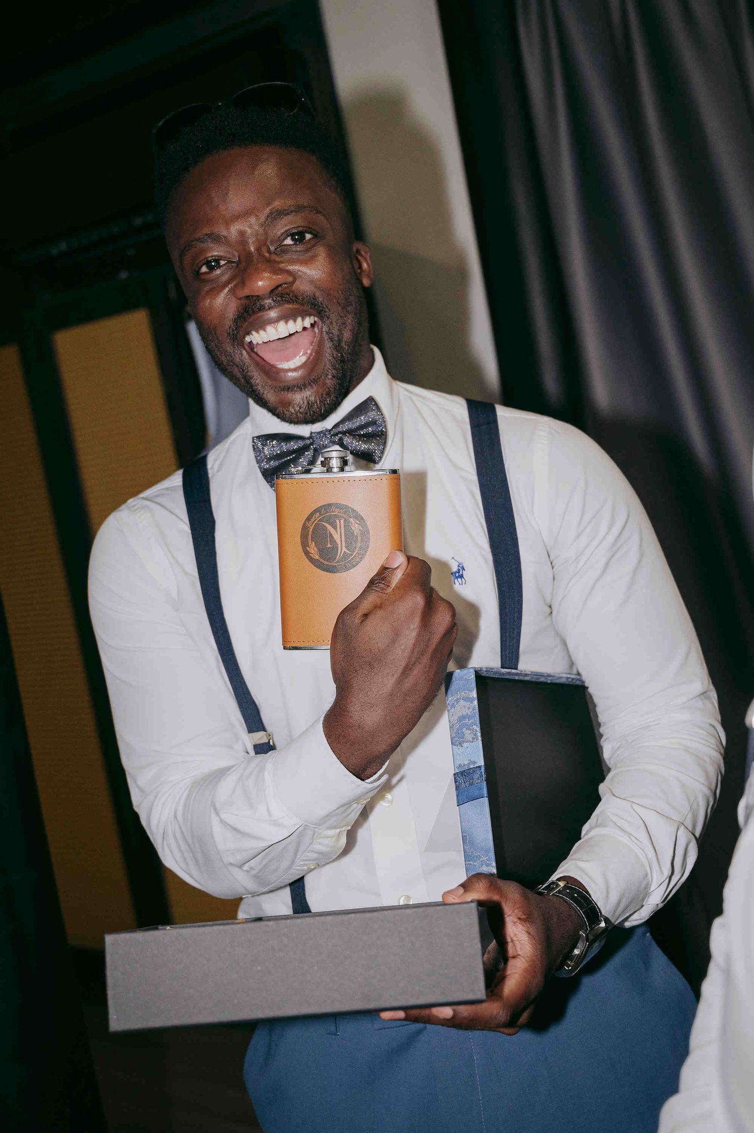 Smiling man in white shirt, bow tie, and suspenders holding a flask and gift box at a celebration.