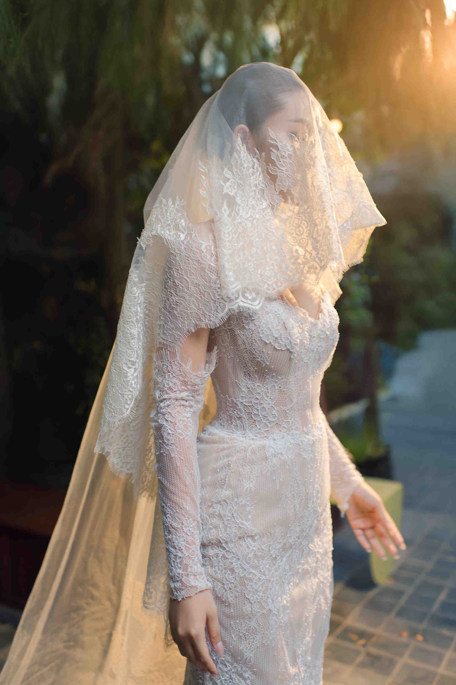 Bride in elegant lace wedding dress and veil covering face, standing outdoors in golden sunset light.