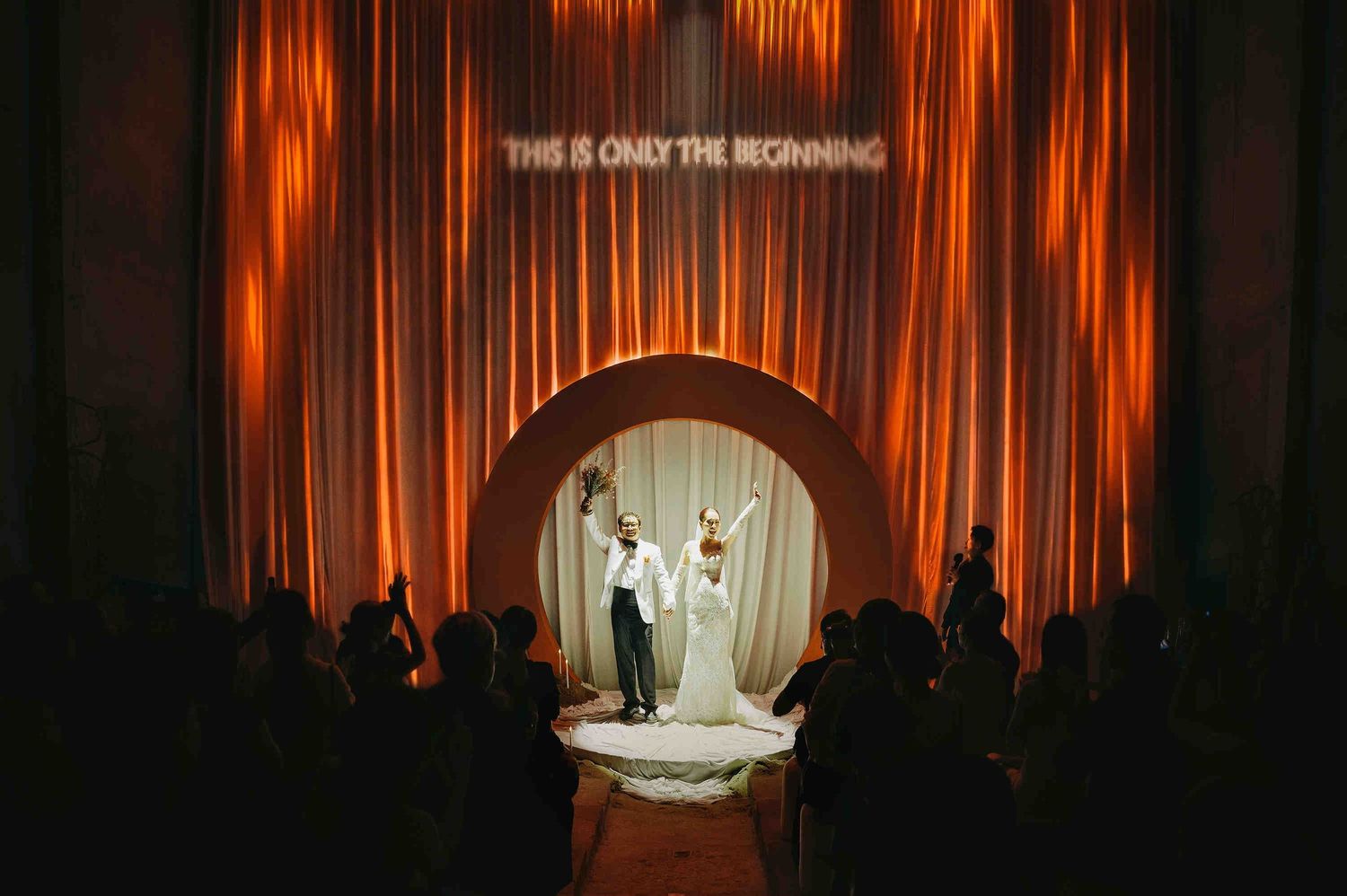 Bride and groom stand beneath glowing arch with 'This Is Only The Beginning' sign at elegant wedding ceremony.