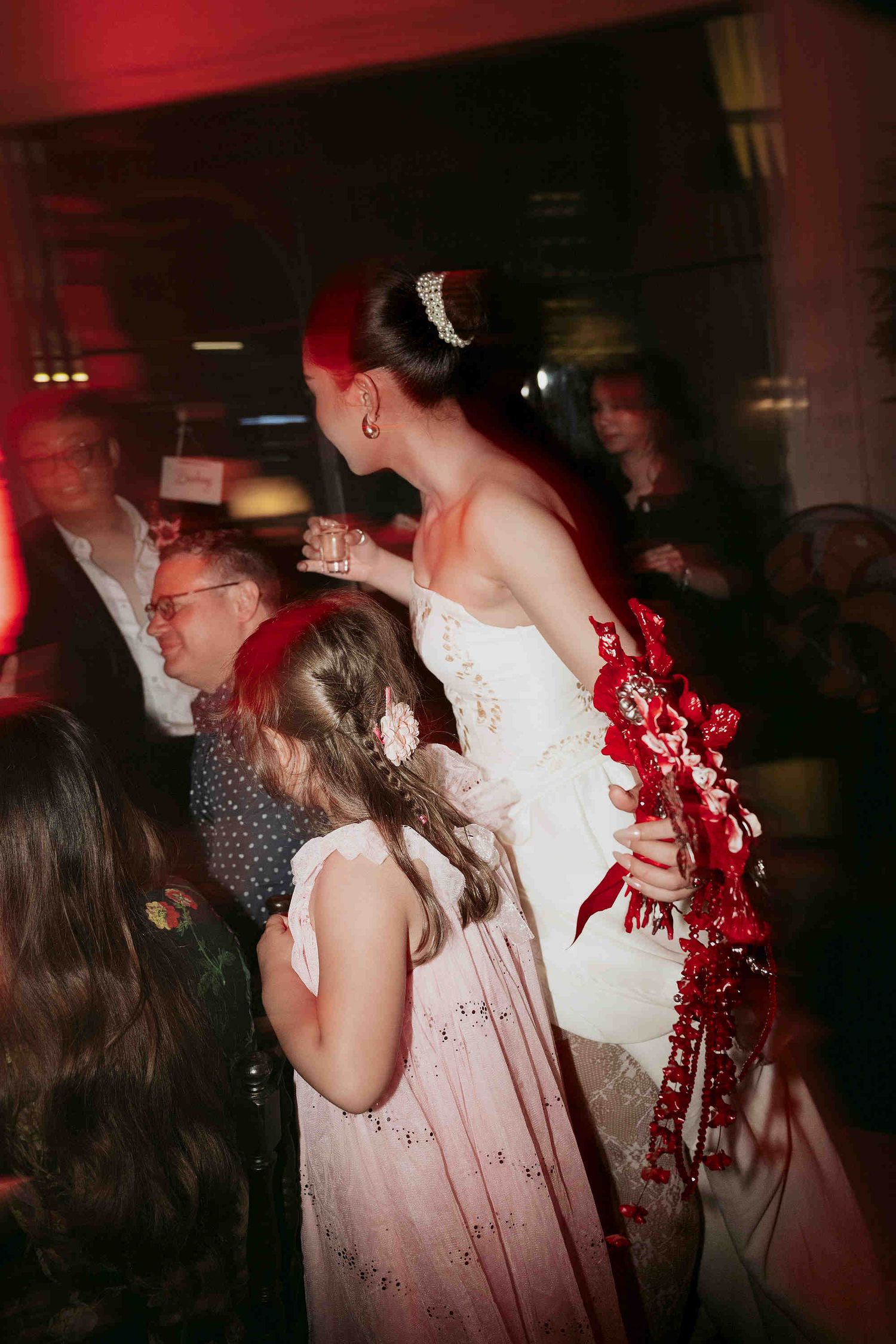 Bride in white gown with red bouquet and hair accessories dancing with flower girl at wedding reception.