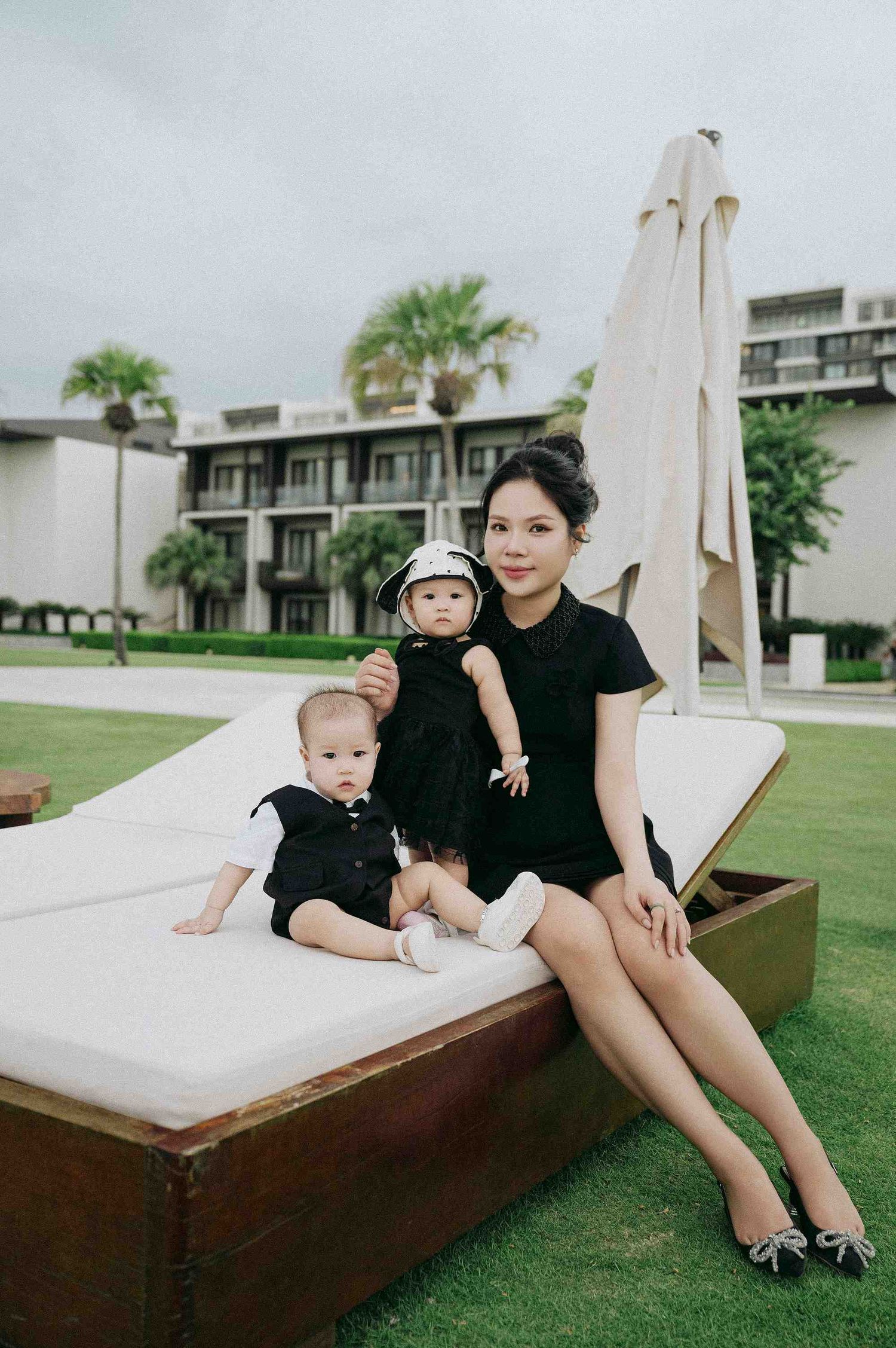 Da Nang family: family posing on an outdoor lounger with palm trees and resort buildings in background, by AnhVuWedding.