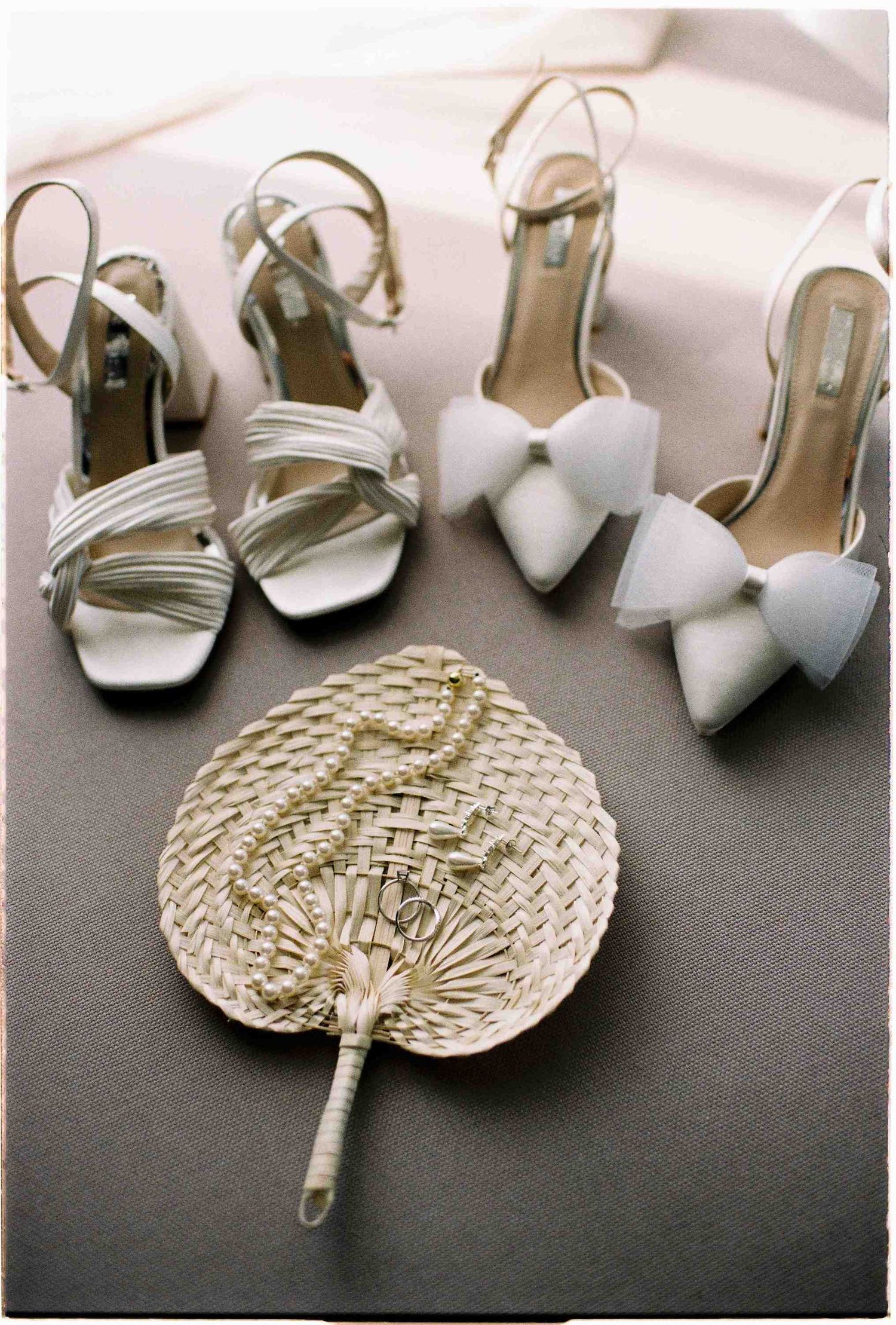 White bridal heels with bow detail and strappy sandals displayed beside a woven straw fan on a neutral surface.
