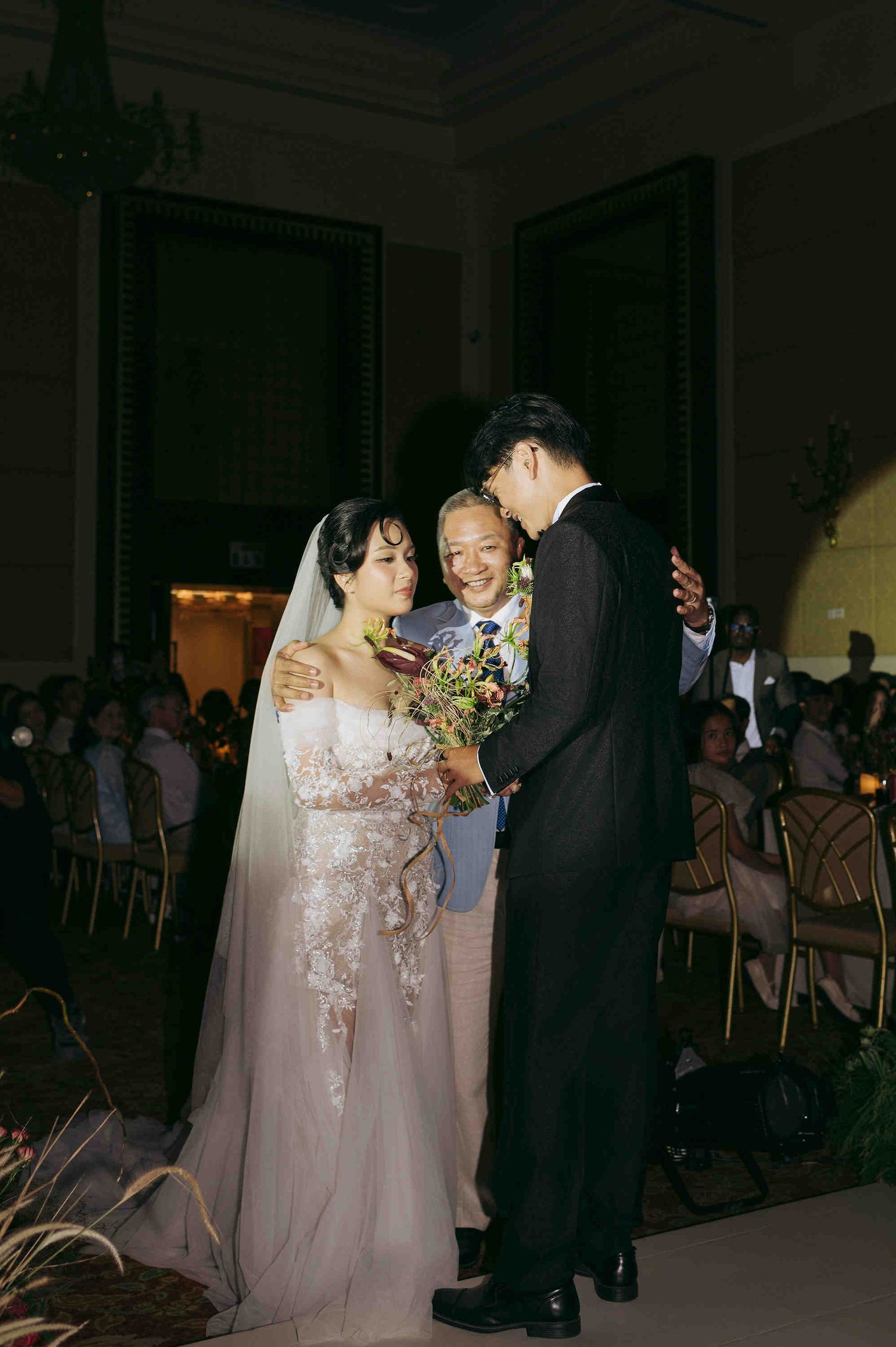 Bride in lace gown and veil receives blessing from elder during indoor wedding ceremony with guests seated.