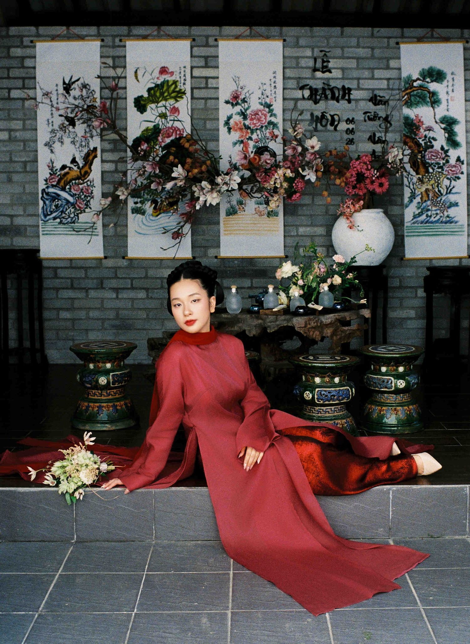 Woman in red ao dai dress poses gracefully in traditional Vietnamese courtyard with floral paintings backdrop.